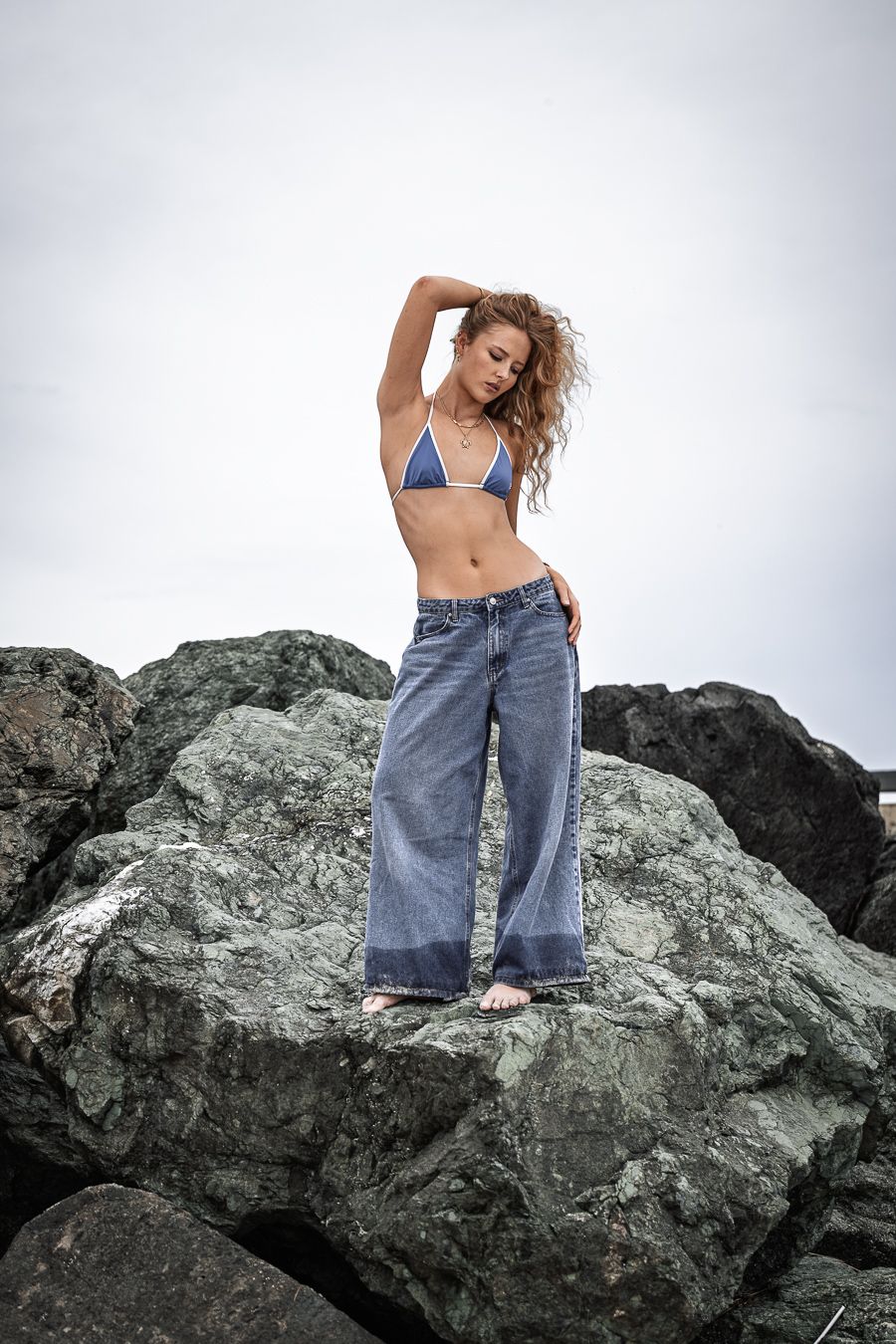 Person in denim jeans and blue top posing artistically on large coastal rocks.
