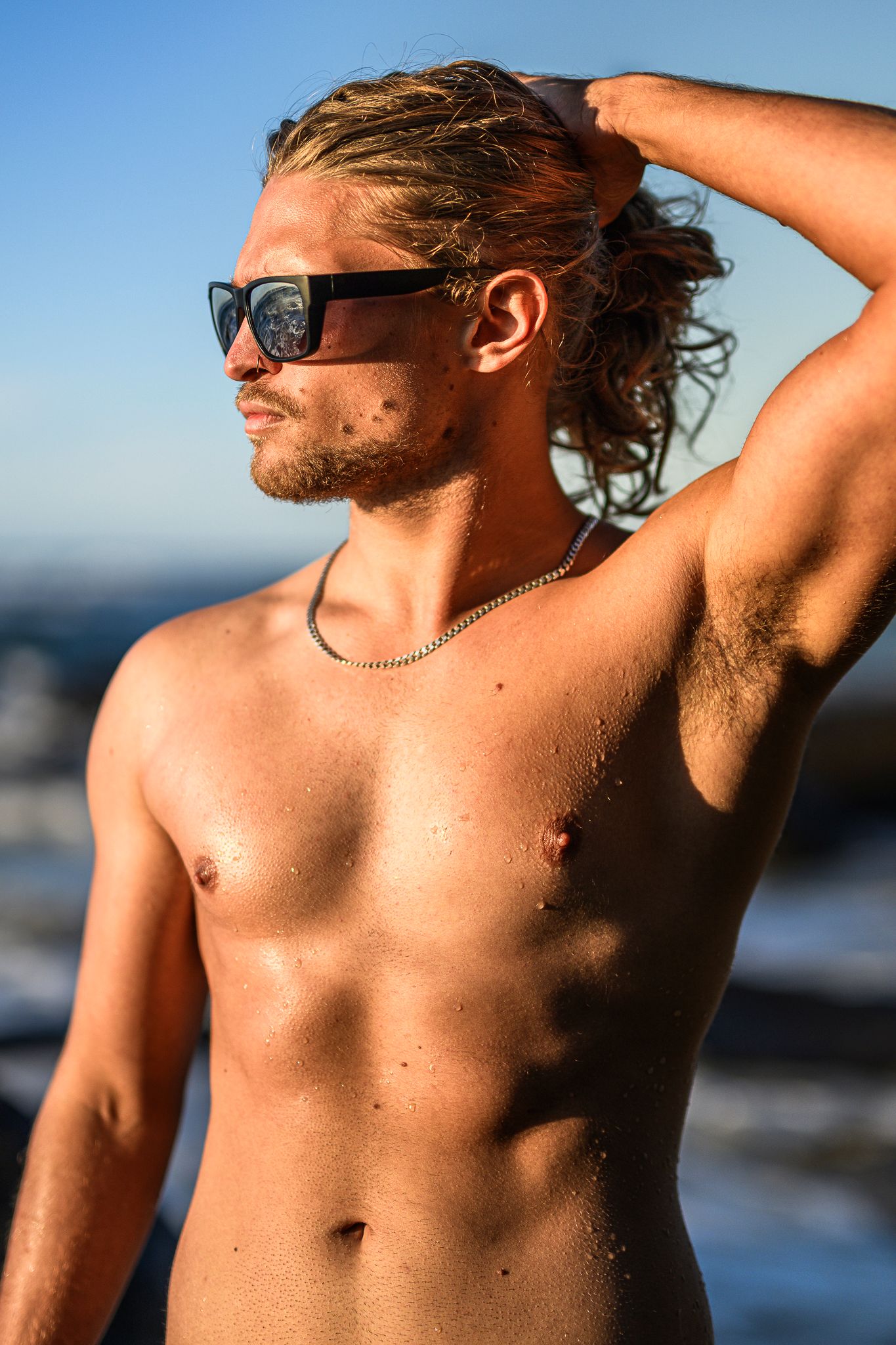 Shirtless person posing on beach at sunset with sunglasses and natural lighting.