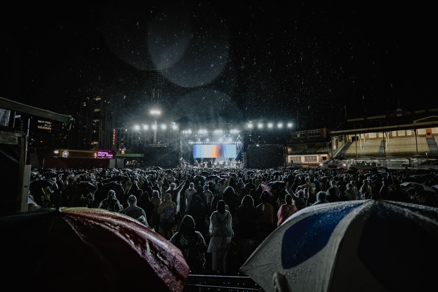 Silhouetted crowd at an outdoor concert venue with bright stage lights illuminating the night sky.