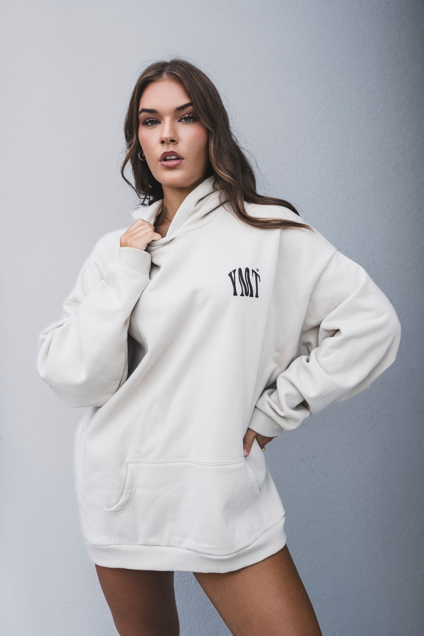 Fashion portrait featuring oversized white hoodie with minimal branding against grey background.