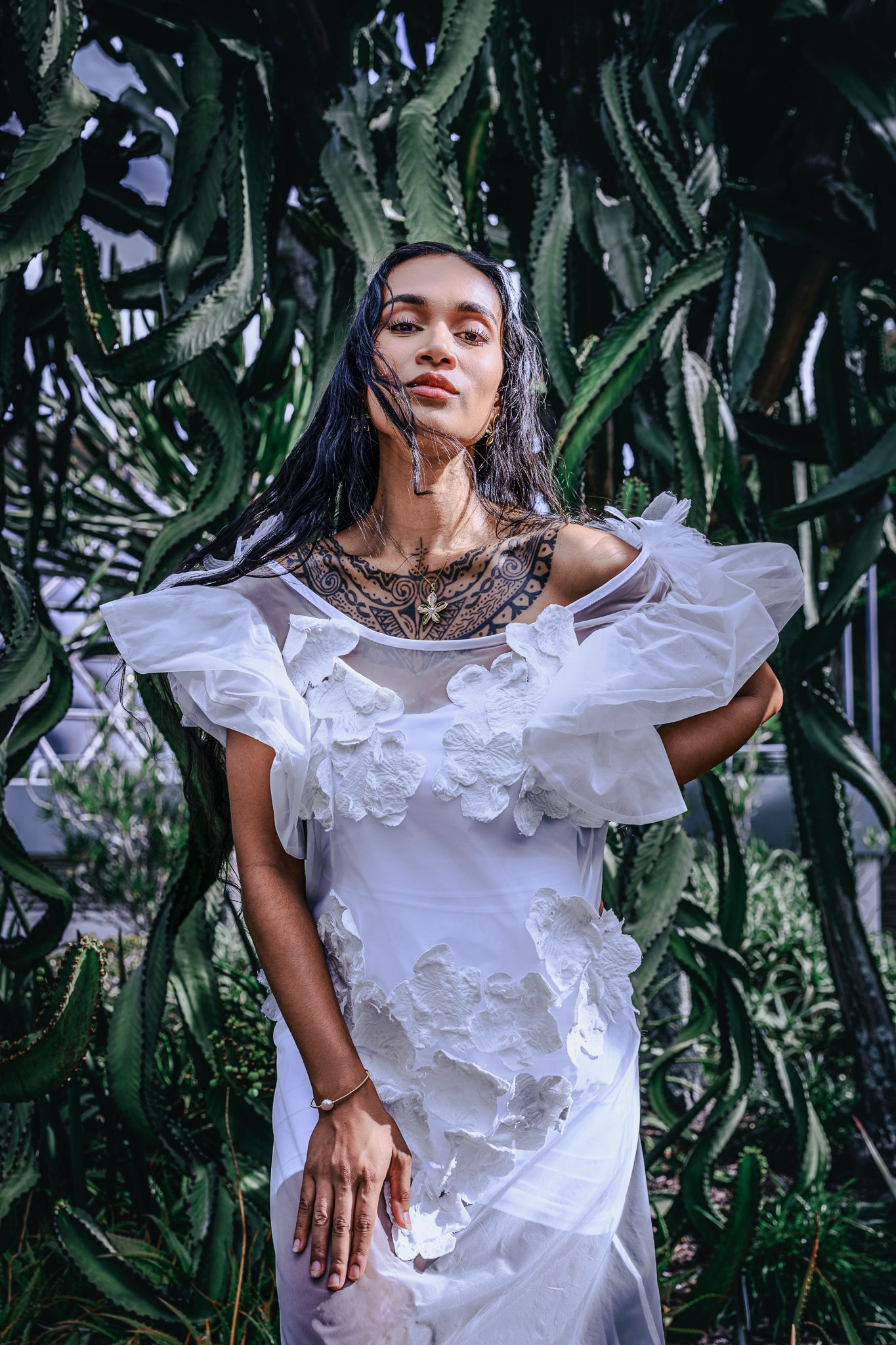 Elegant woman in white ruffled floral dress with gold necklace posing against towering green cactus backdrop.