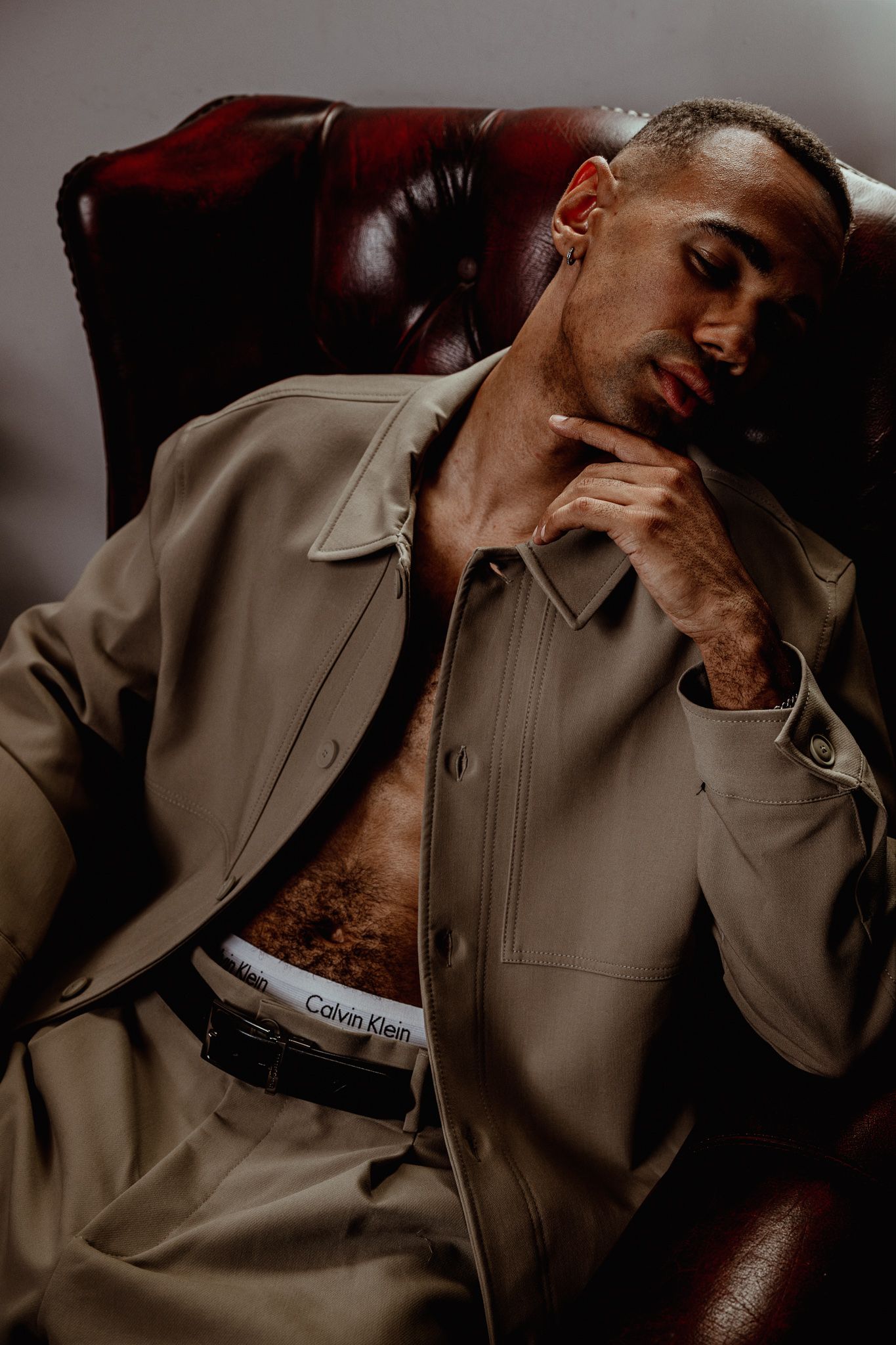 Man in beige jacket and Calvin Klein underwear sitting in leather chair with hand on chin.