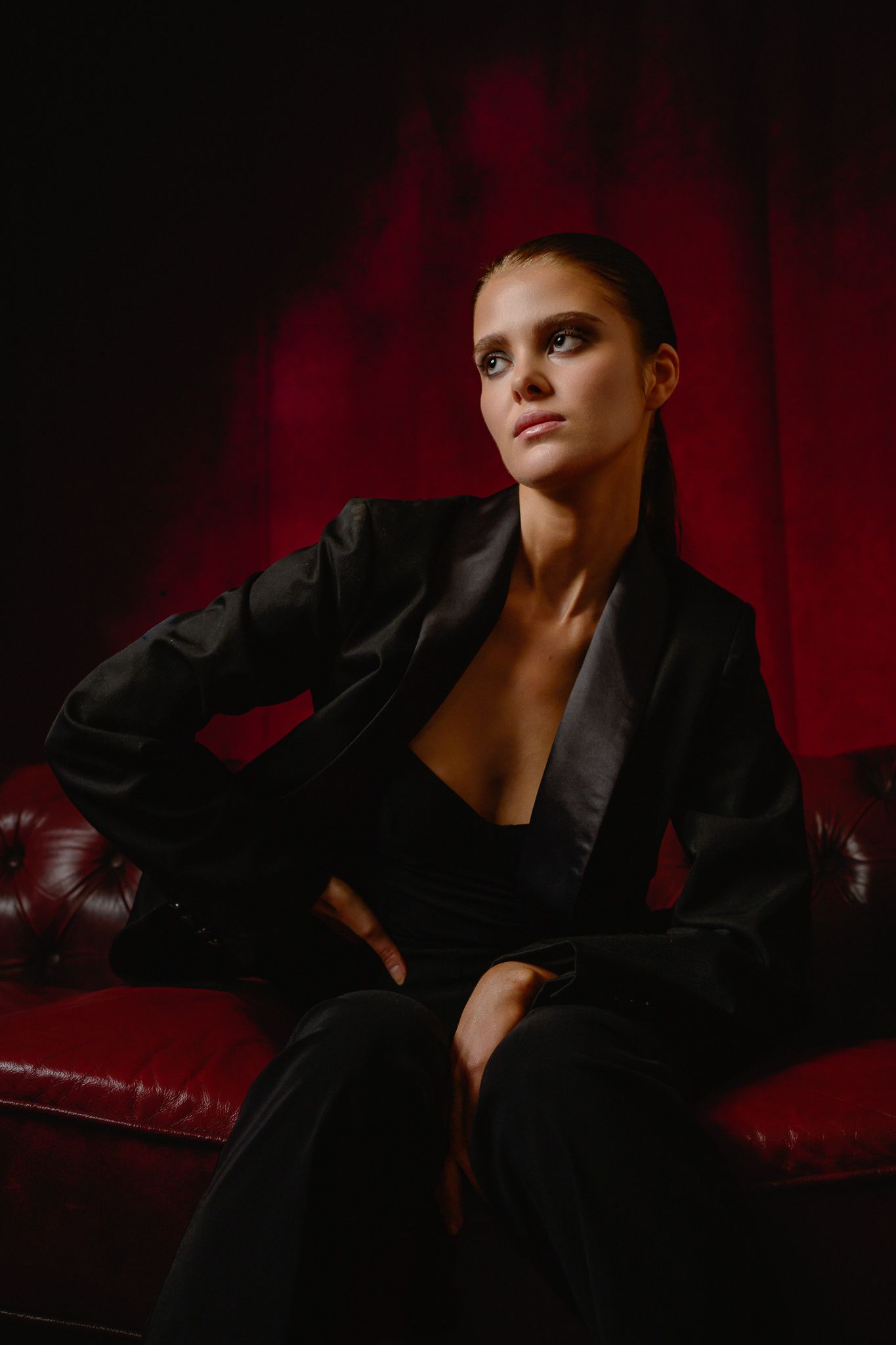Woman in black blazer suit seated on red leather sofa against dramatic red backdrop in moody fashion portrait.