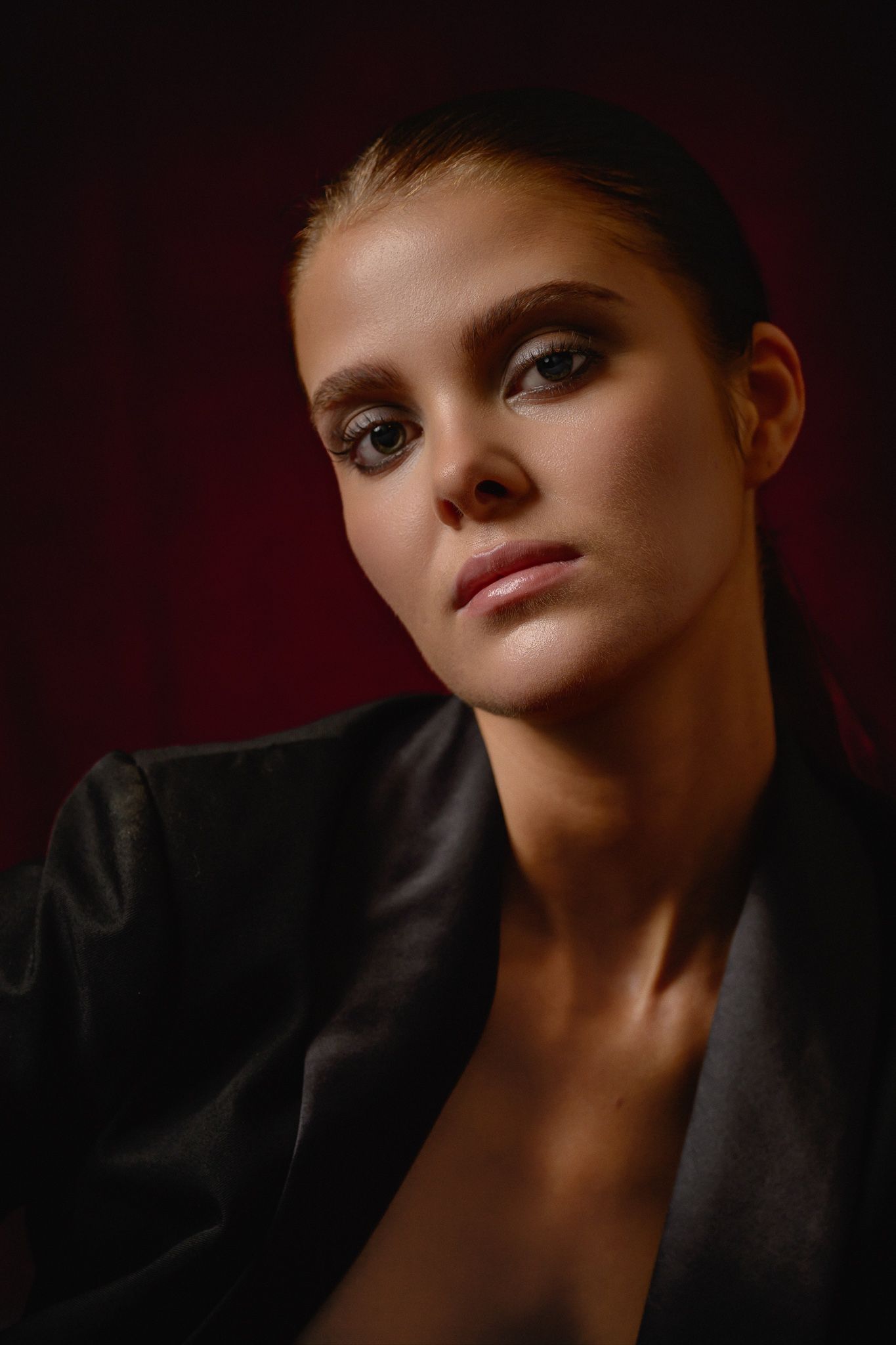 A young woman in a black blazer poses against a dark red background with dramatic makeup and a confident gaze.