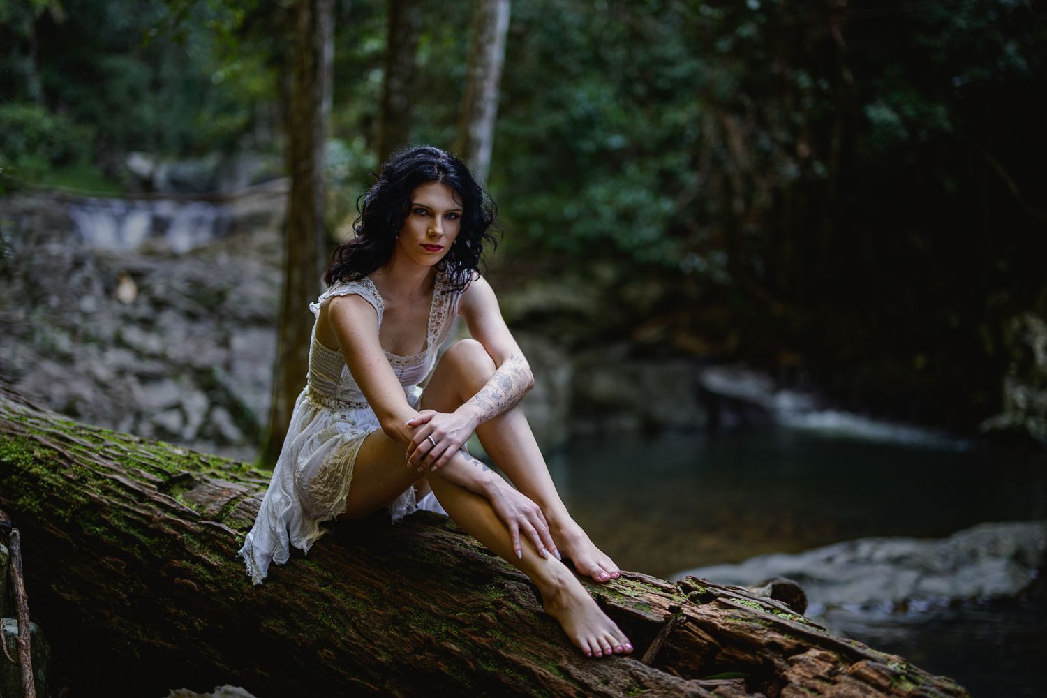 Woman in lace dress sitting on mossy log by forest stream, surrounded by lush green trees and rocks.