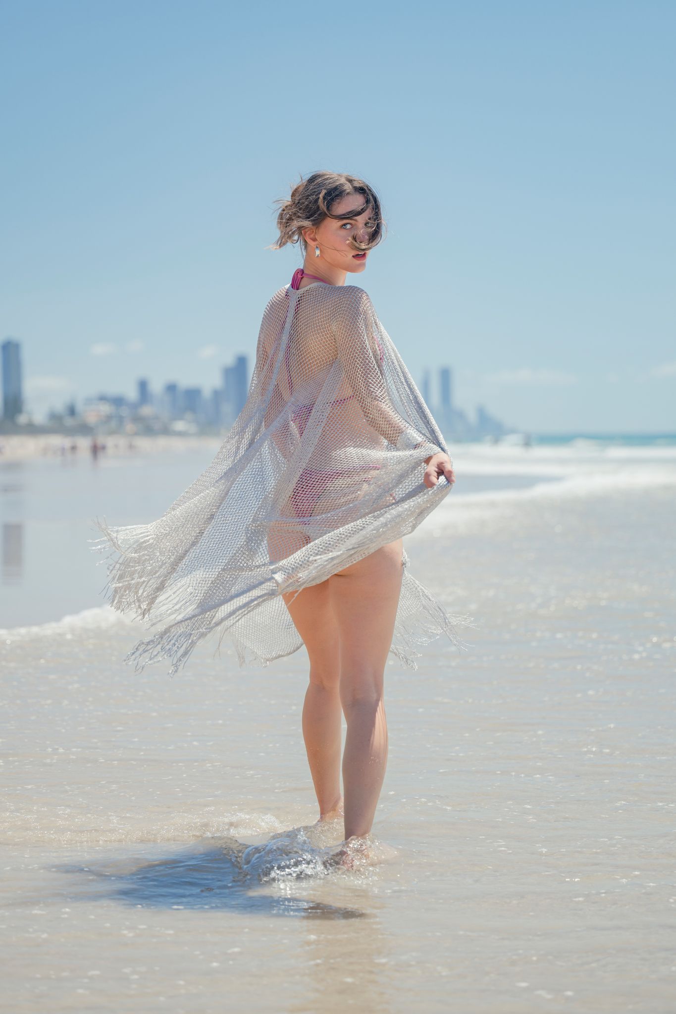 Model in sheer cover-up poses on beach with city skyline in background.