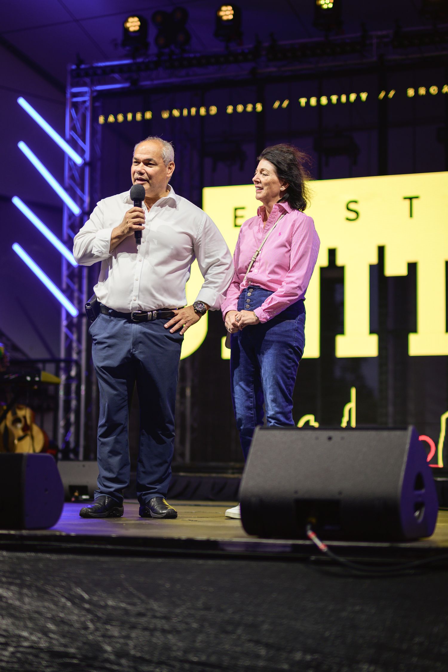 Two speakers presenting on stage with colorful lighting and event signage in the background.