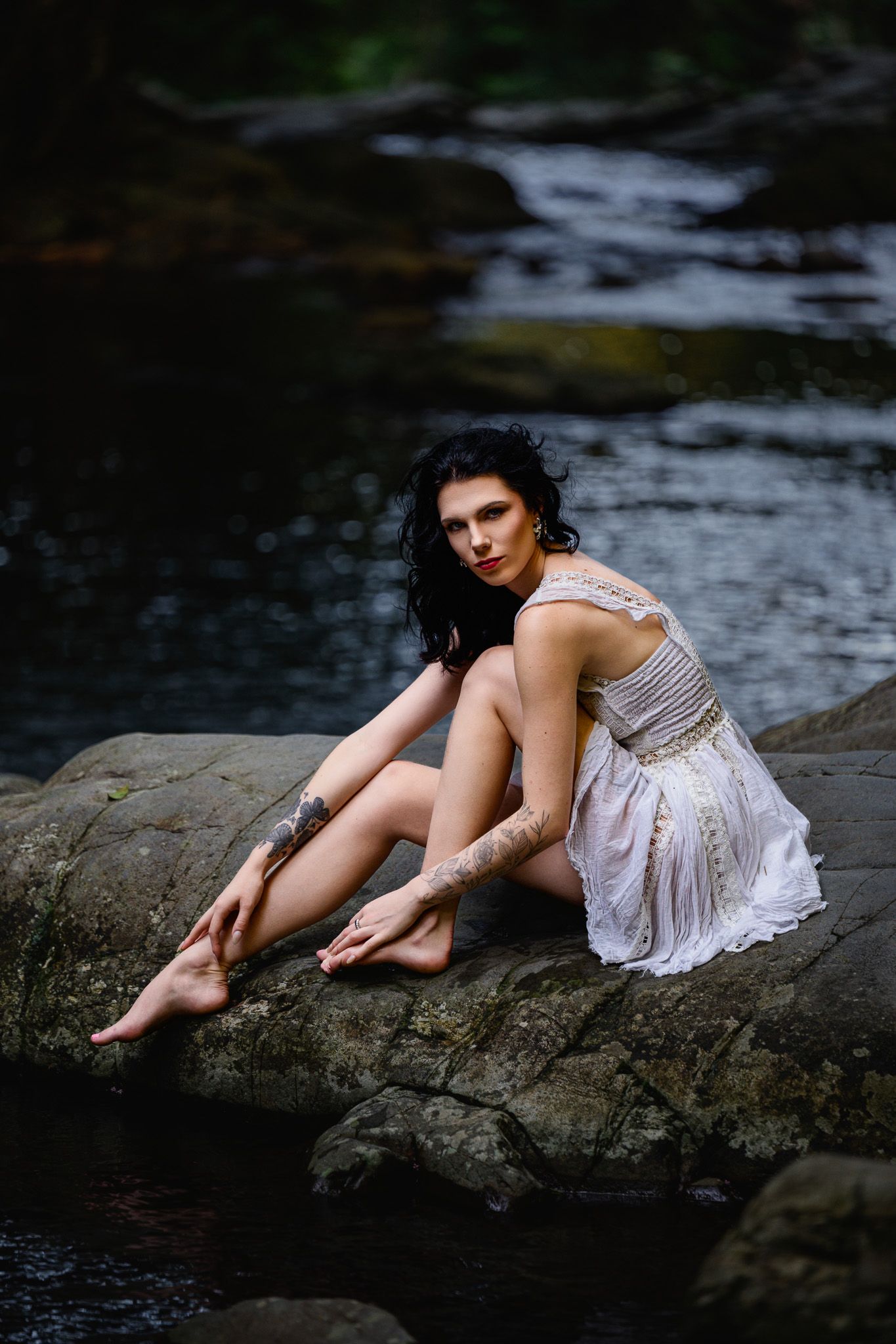 Dark-haired woman in white dress sitting barefoot on rocks by a forest stream, with tattoos on her arms.