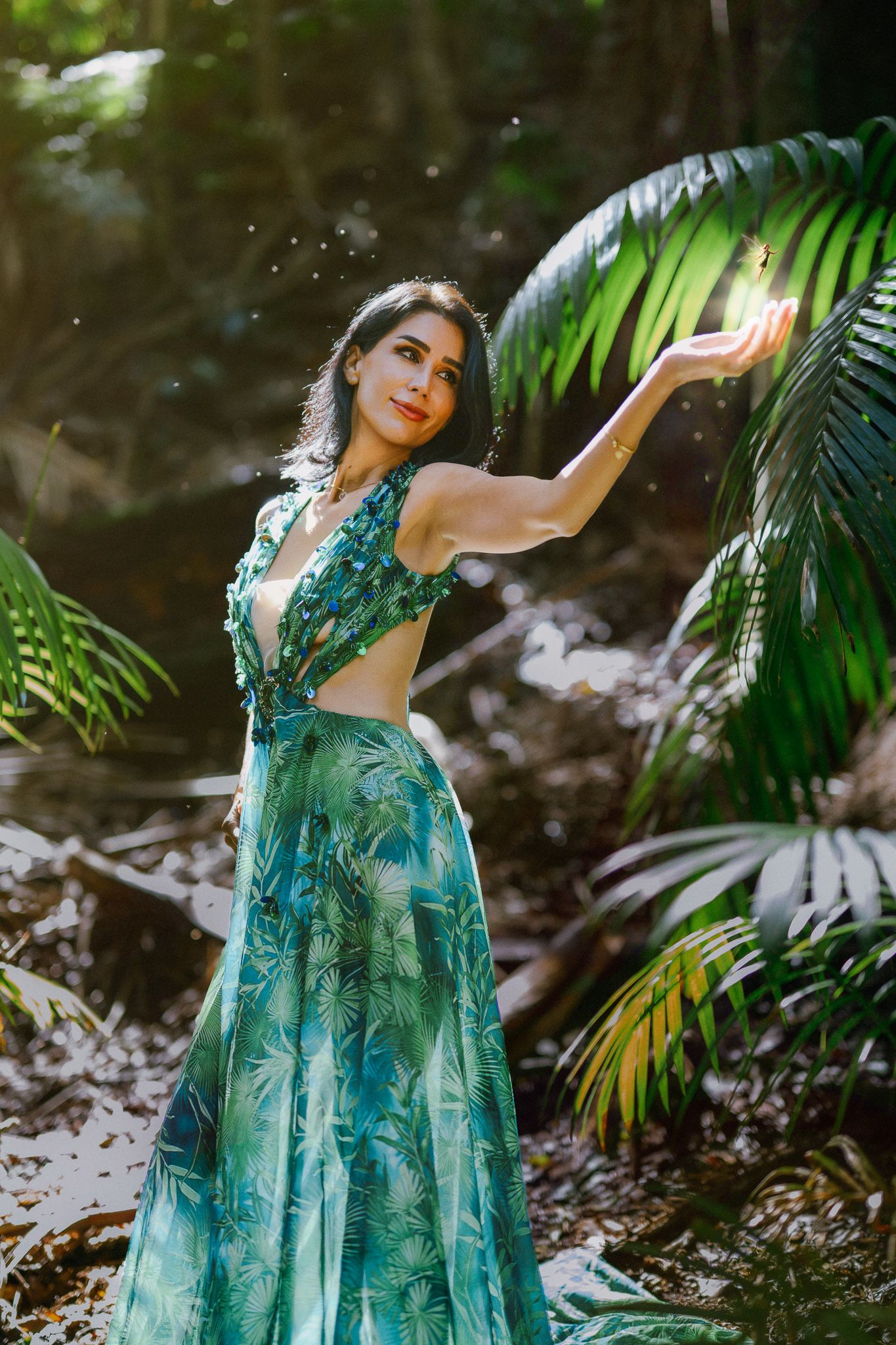 Woman in teal tropical print dress poses gracefully among lush palm leaves in a sunlit forest setting.