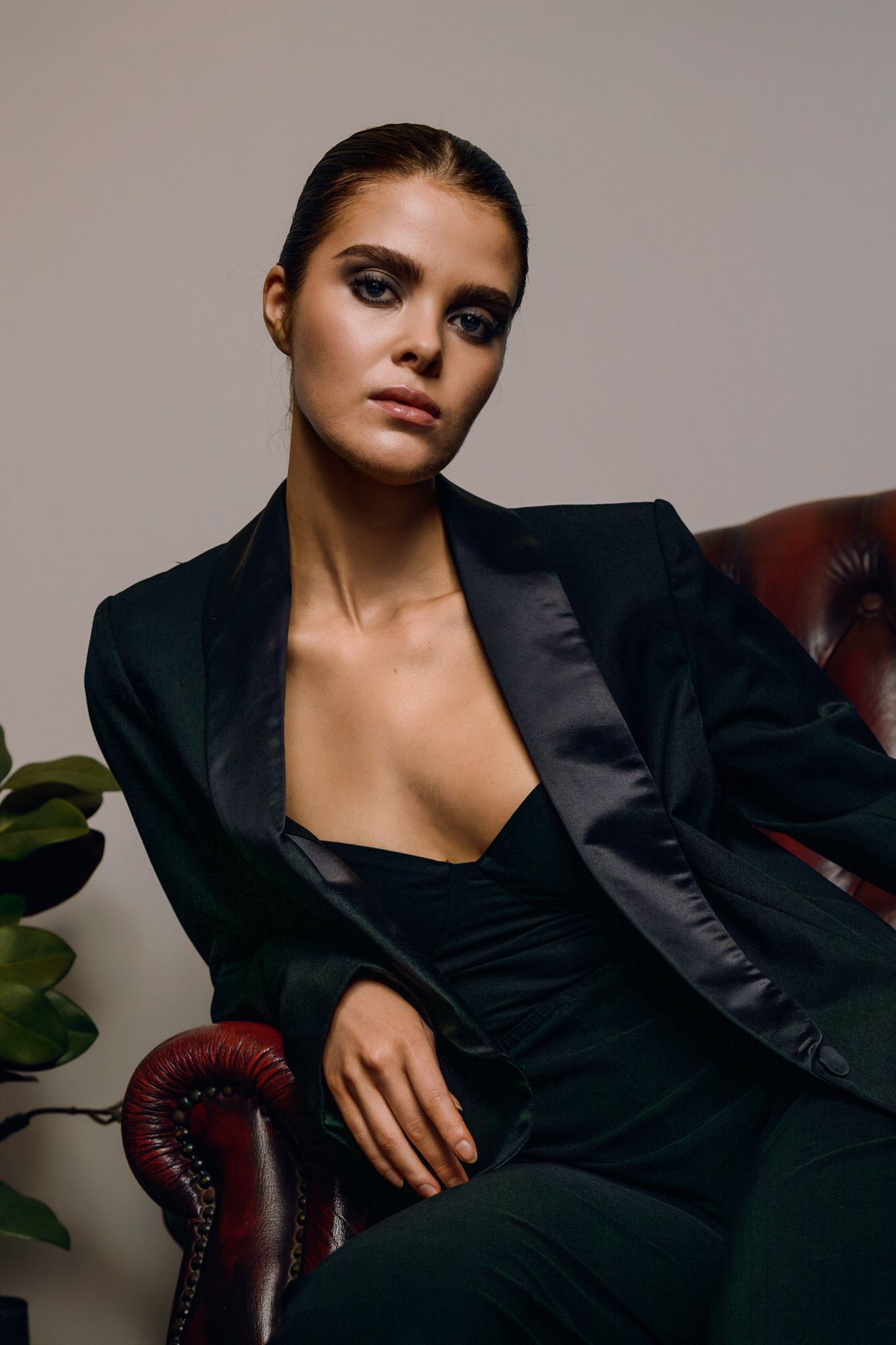 Elegant woman in black blazer suit seated in leather chair with dramatic smoky eye makeup.