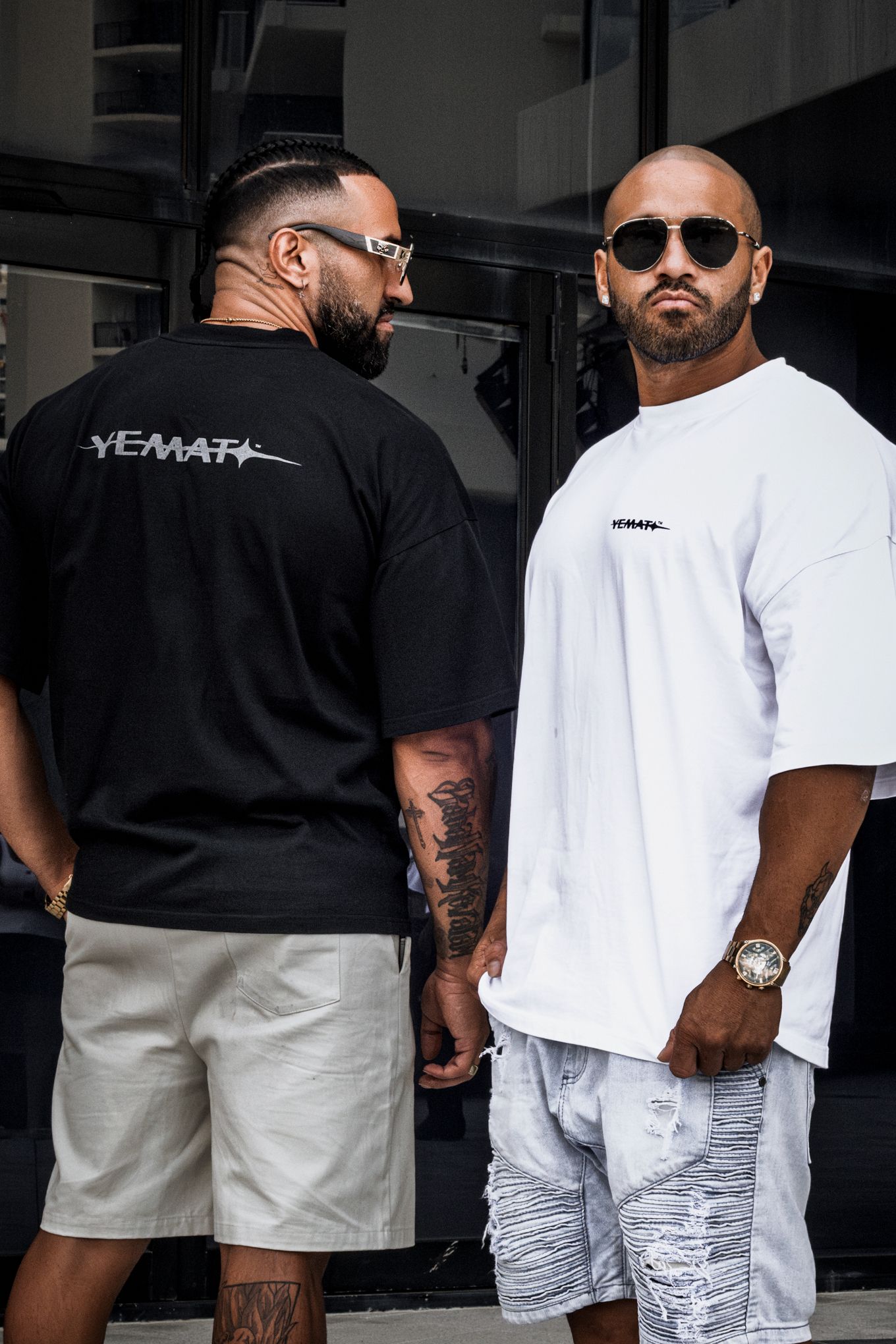 Two men wearing Yemat branded oversized t-shirts in black and white with shorts and sunglasses pose outdoors.
