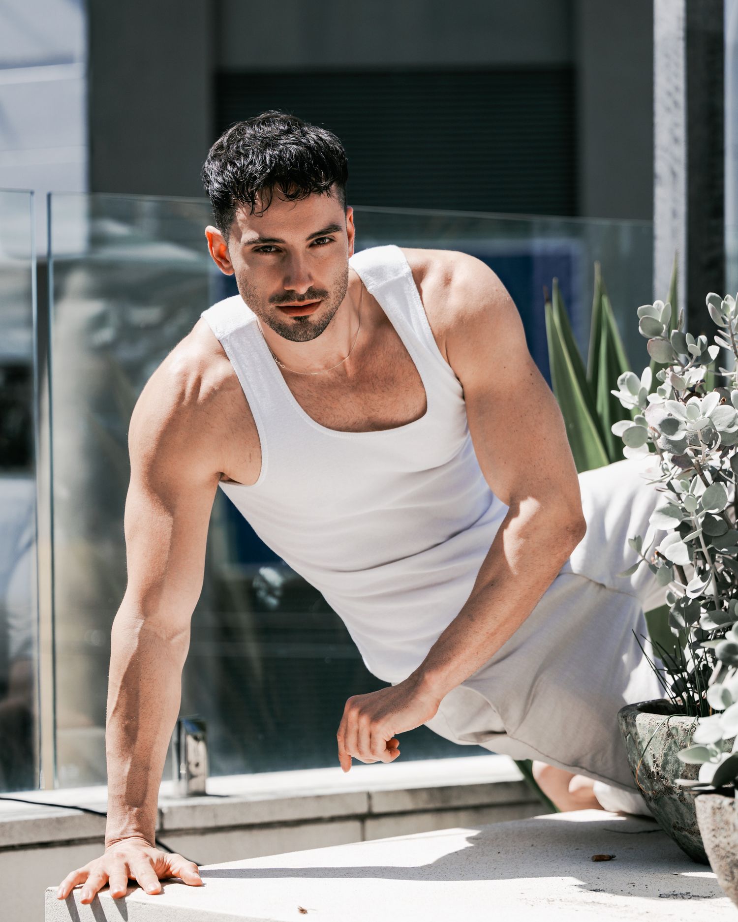 Athletic man in white tank top leaning on windowsill with decorative plants.
