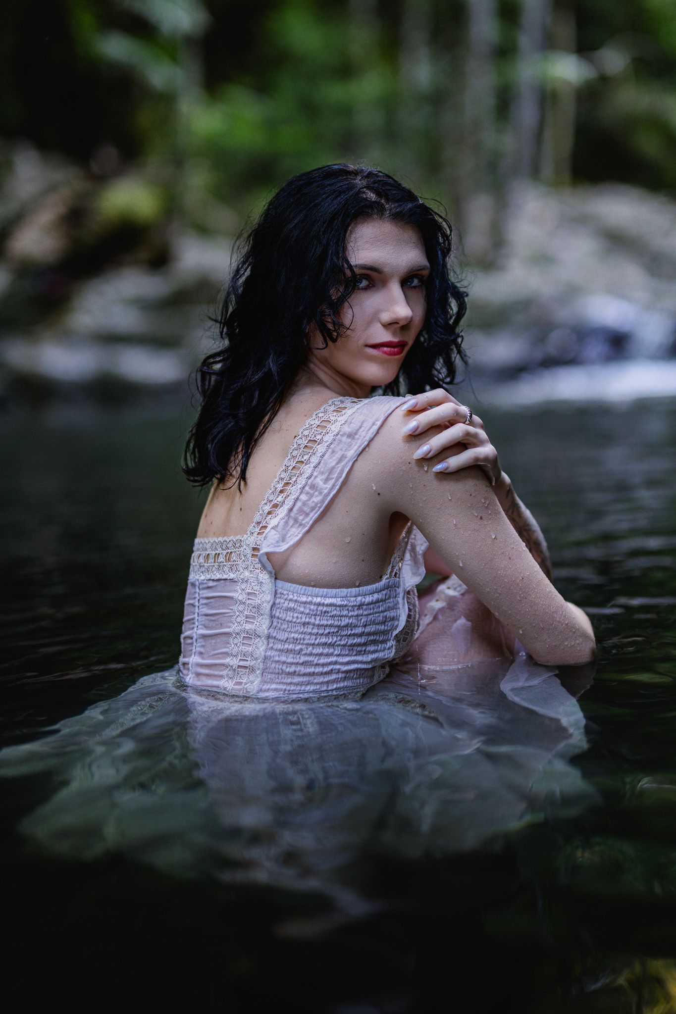 Dark-haired woman in white lace dress sitting in a forest stream, looking over her shoulder.