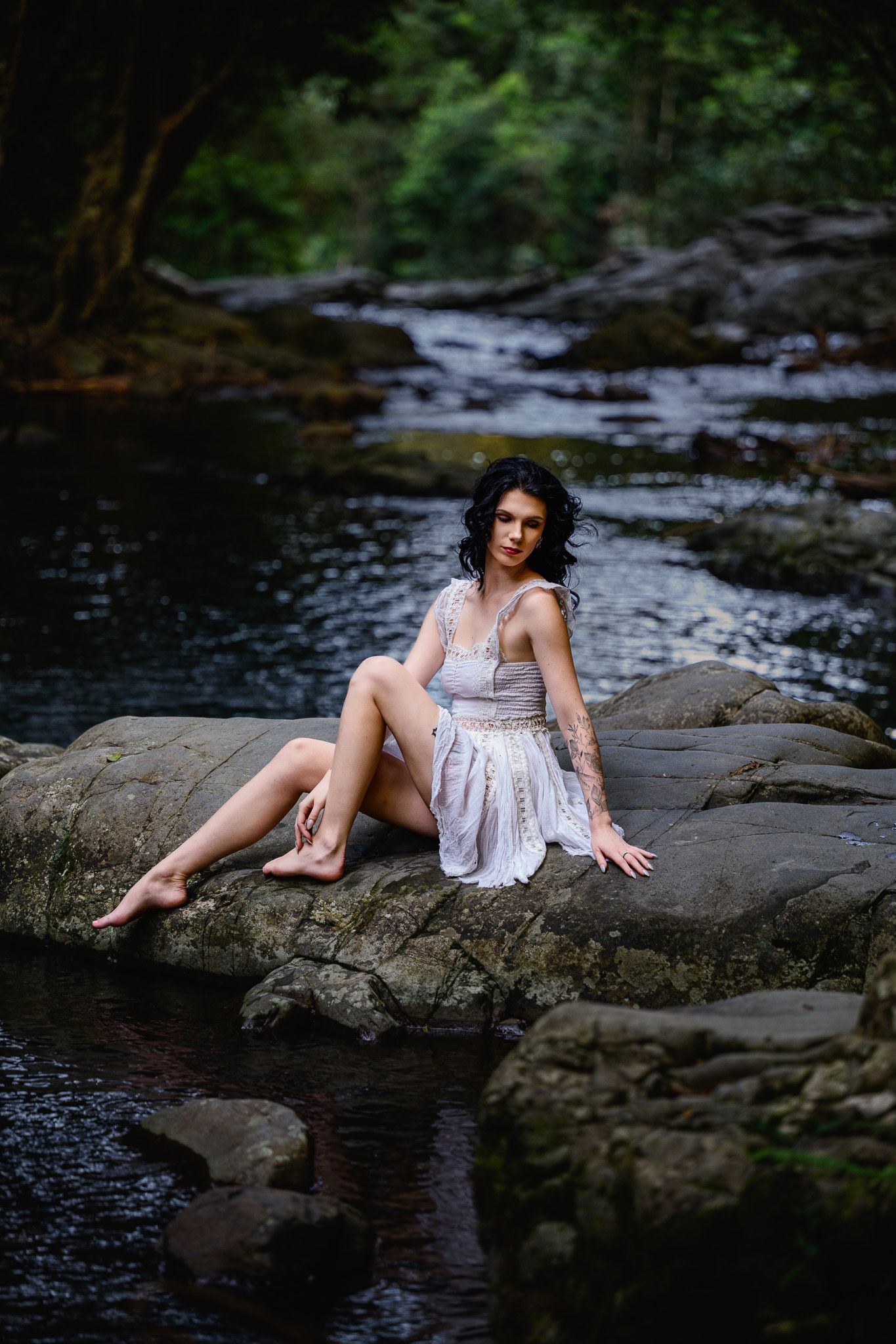 A woman in a white dress sits barefoot on rocks by a forest stream, posing gracefully in a serene natural setting.