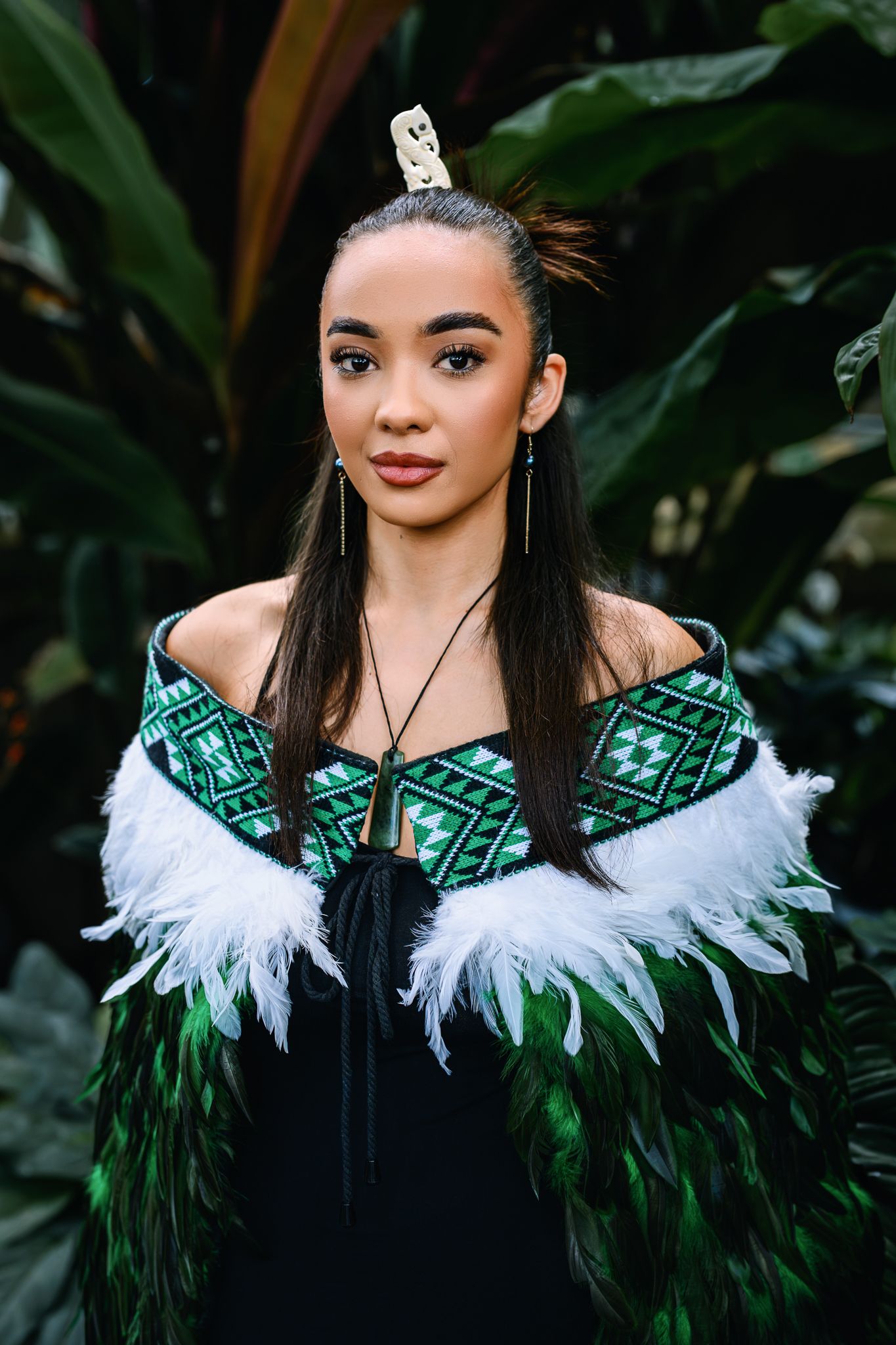 Young woman in traditional Maori attire with green feather cloak, bone pendant, and hair ornament, posed before tropical foliage.