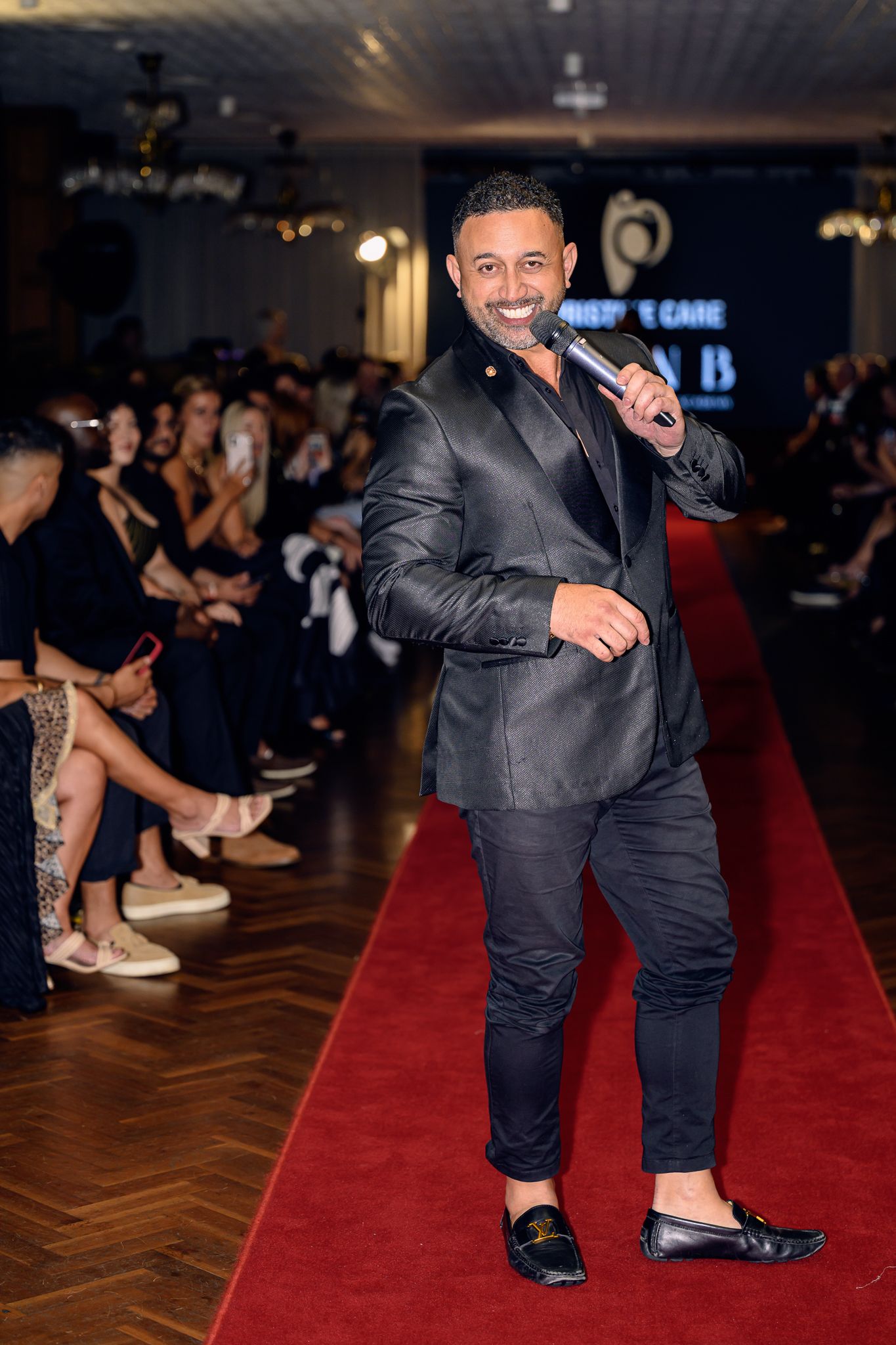 A performer in a dark suit holds a microphone while walking down a red carpet runway.