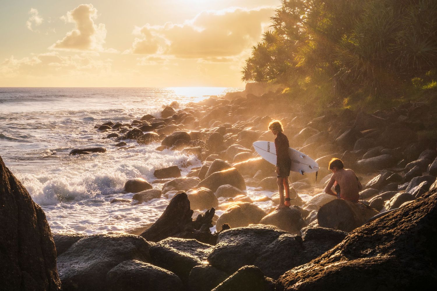 Two surfers on rocky shoreline at golden sunset with ocean waves and lush tropical trees in background.