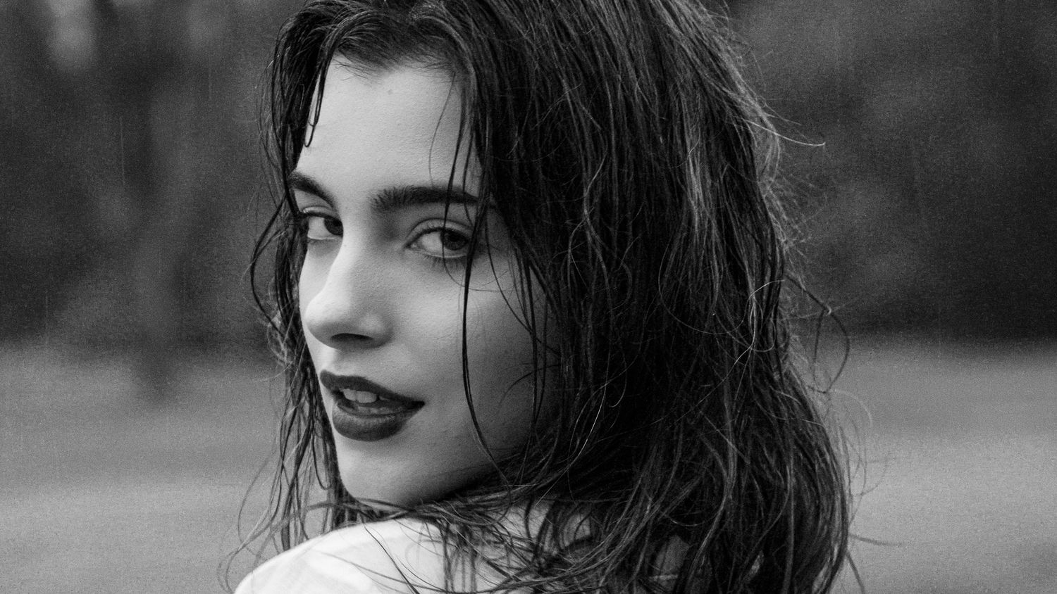Black and white portrait of a young woman with dark wavy hair and bold lips, gazing over her shoulder.