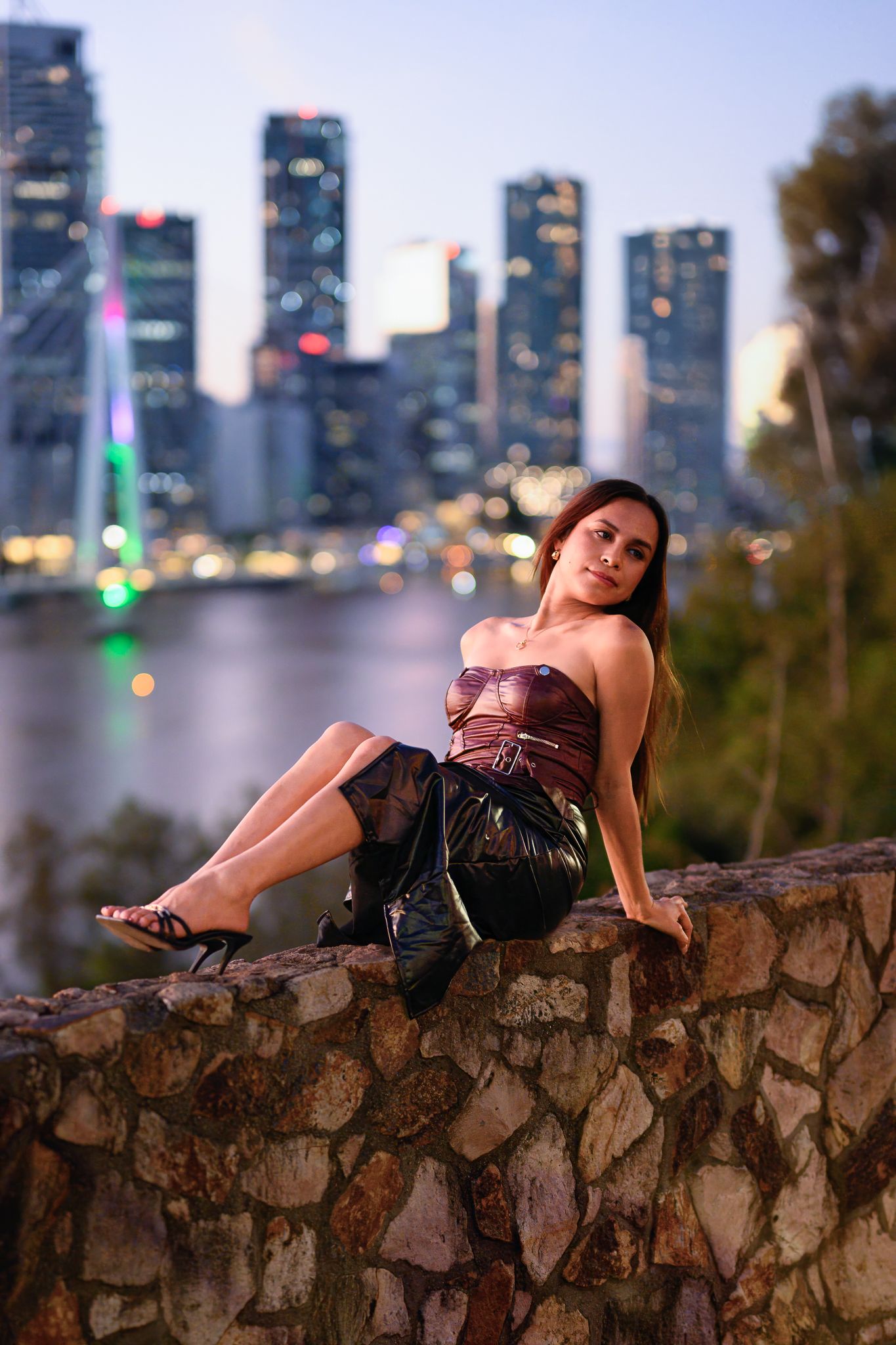 Fashion model poses dramatically on stone wall against urban skyline at sunset.