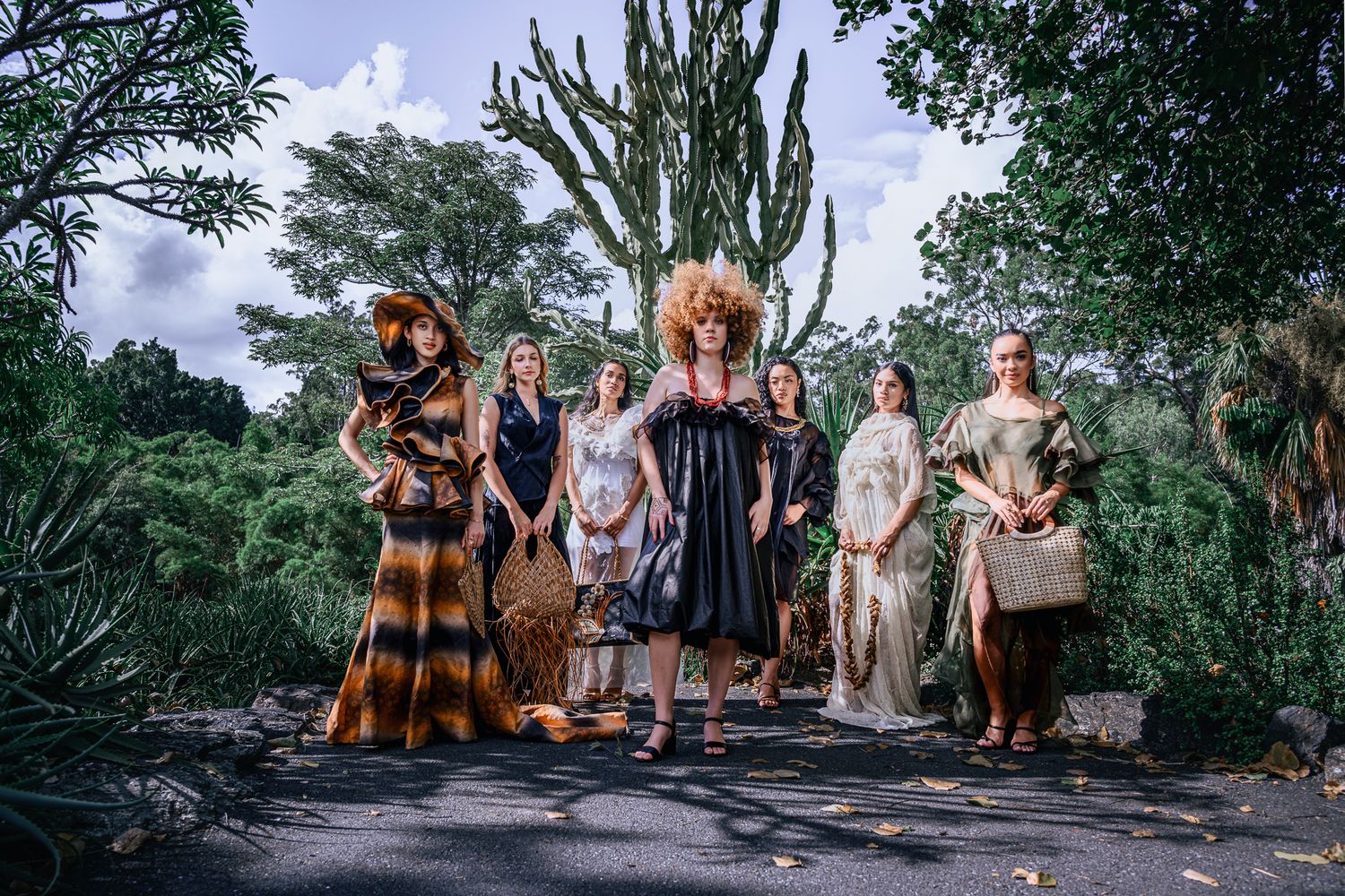 Group of women in bohemian fashion poses outdoors among lush tropical greenery and cacti.