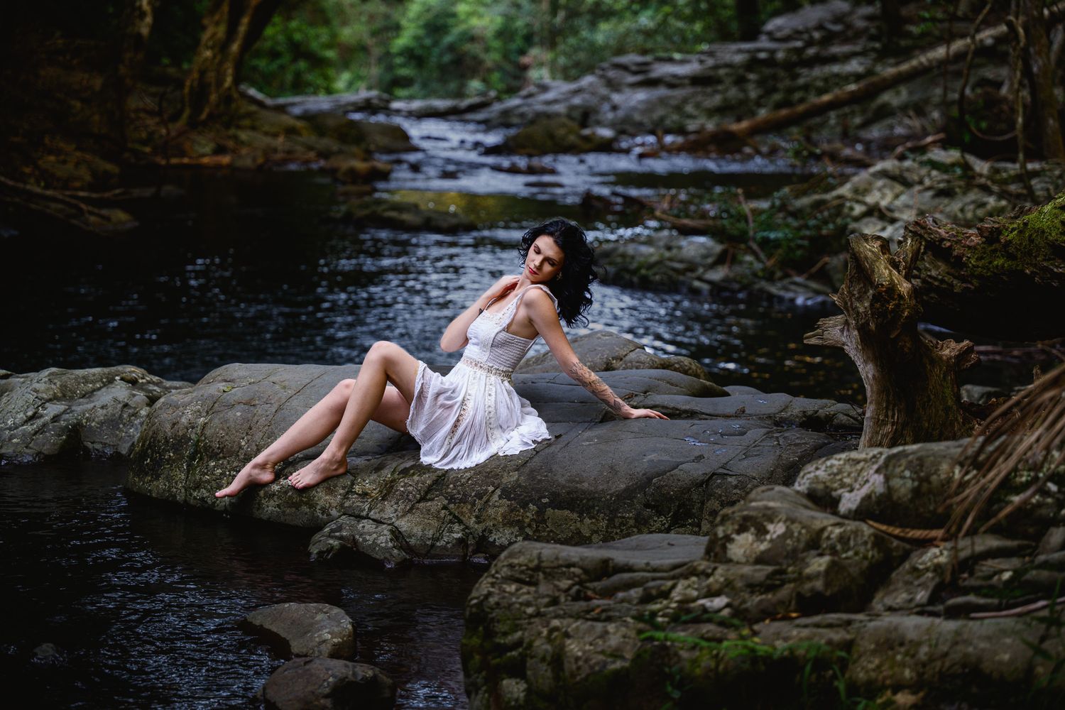 Woman in white dress sitting on rocks by a serene forest stream, posing gracefully in a lush natural setting.