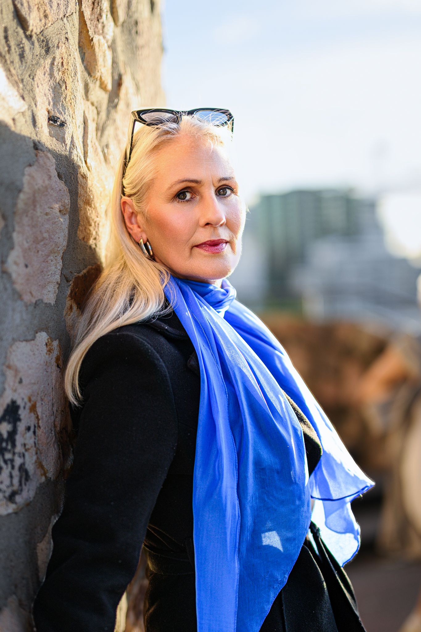 Woman wearing a black coat and bright blue scarf poses against stone wall in outdoor portrait series.