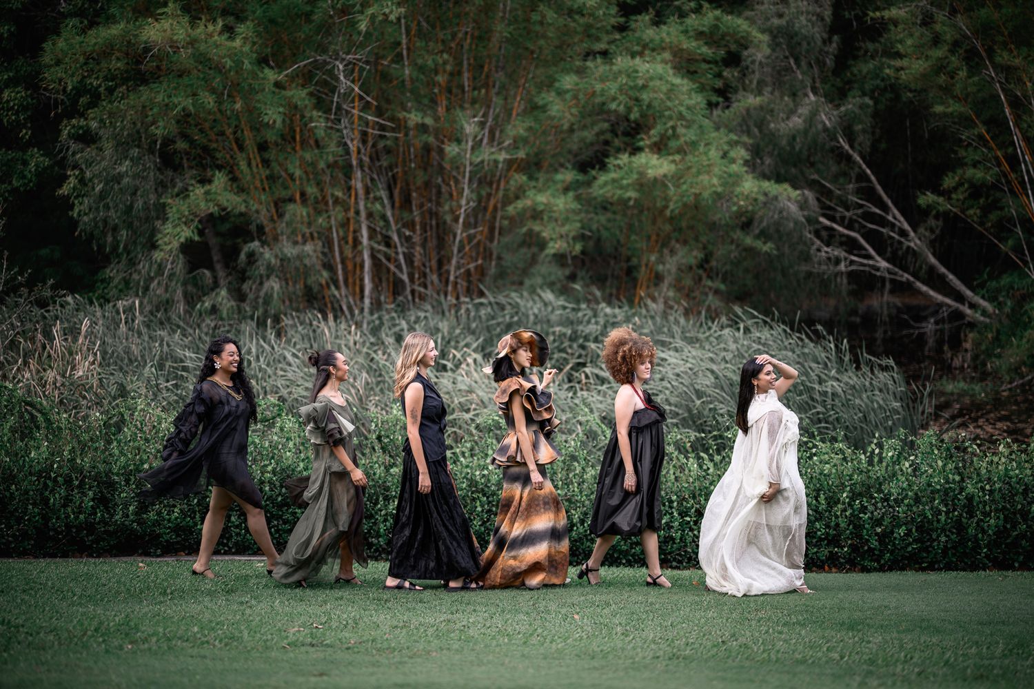 Six women in elegant dresses walking across green lawn with lush trees in background.