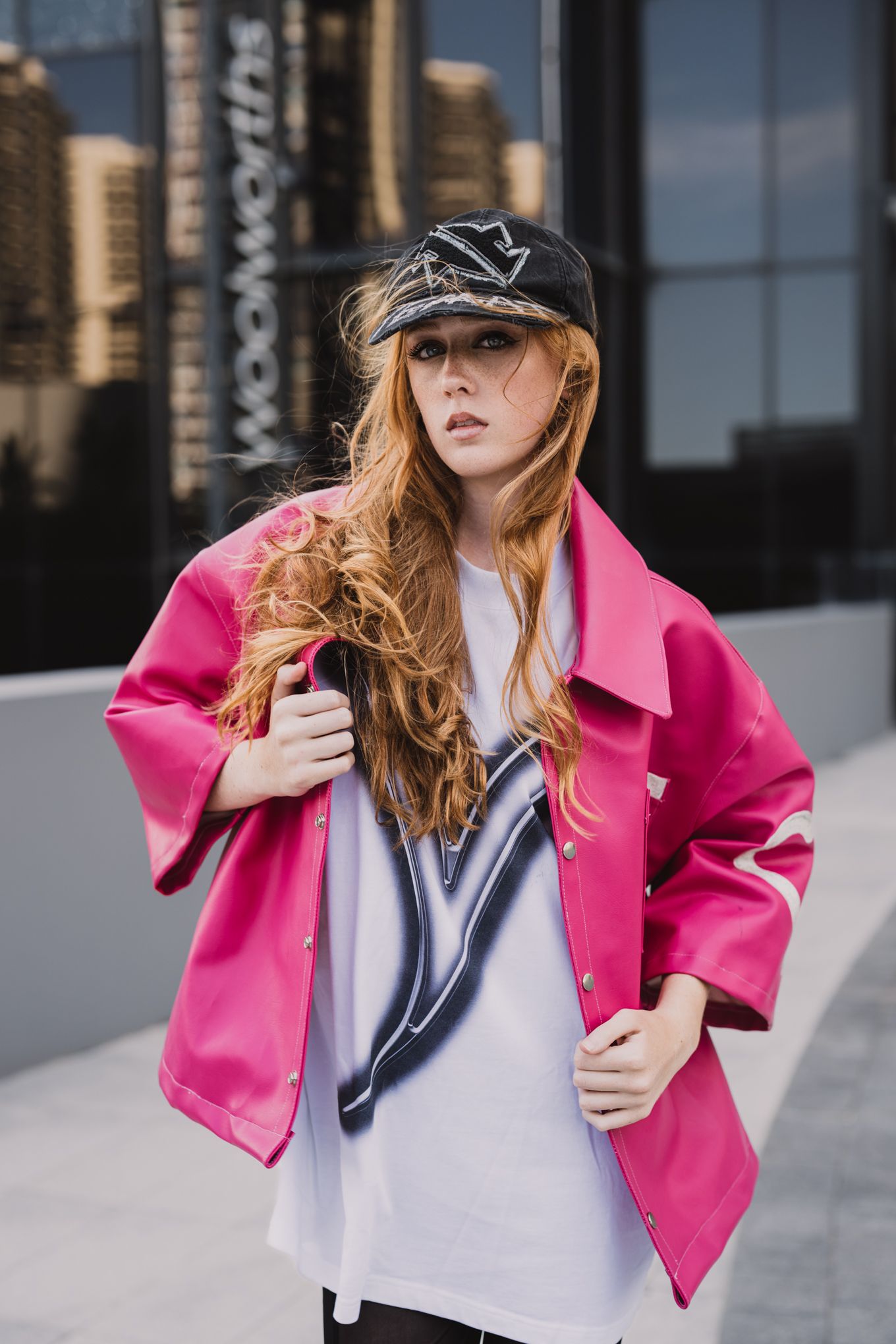 Street style look with bright pink coat and baseball cap against urban backdrop.