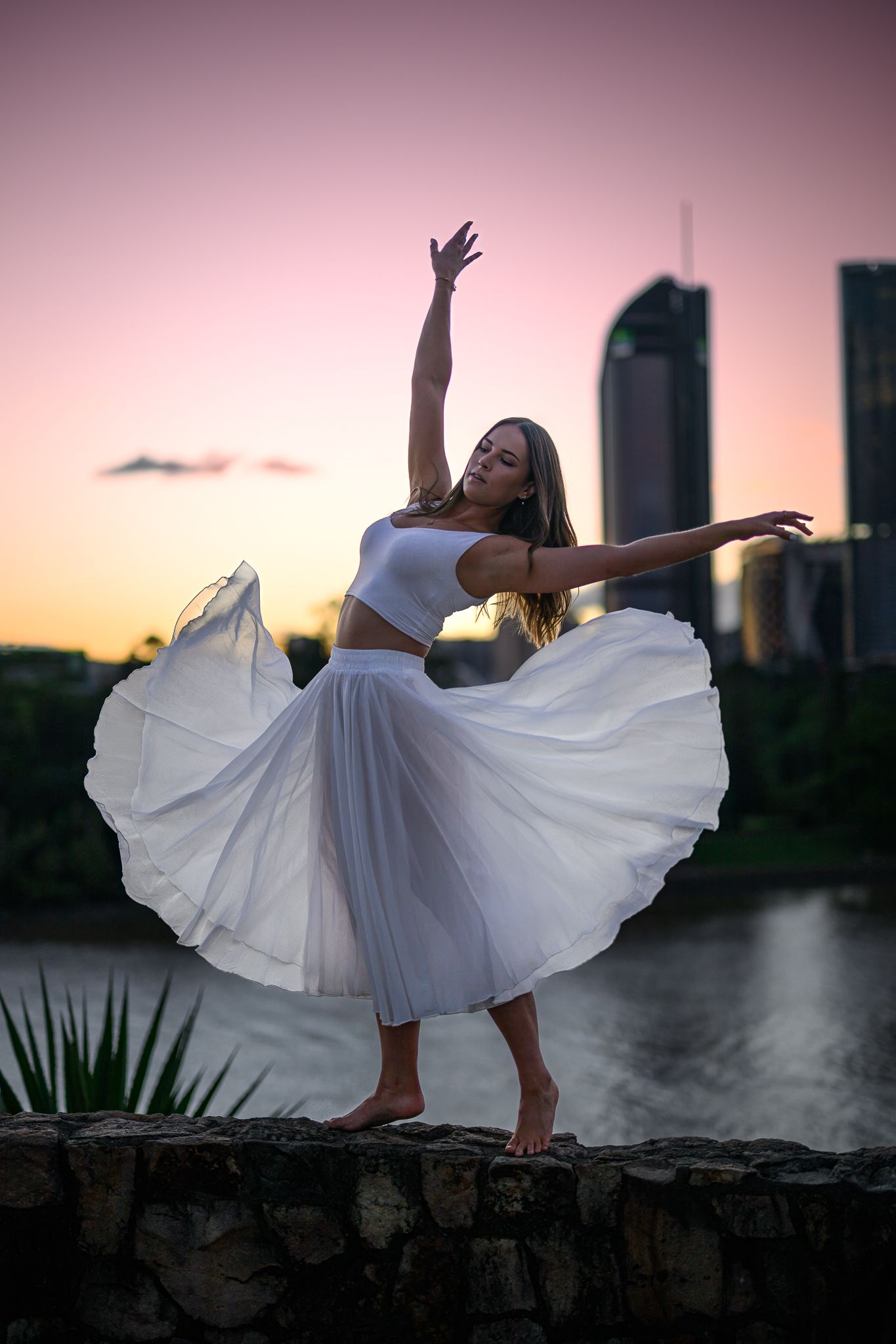 Dancer in flowing white dress performs graceful movements against city backdrop at dusk.
