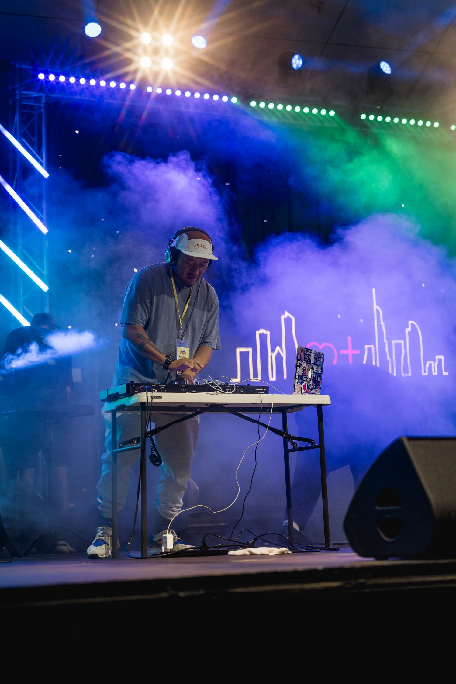 DJ performing at mixing table with cityscape backdrop and colorful stage lighting.