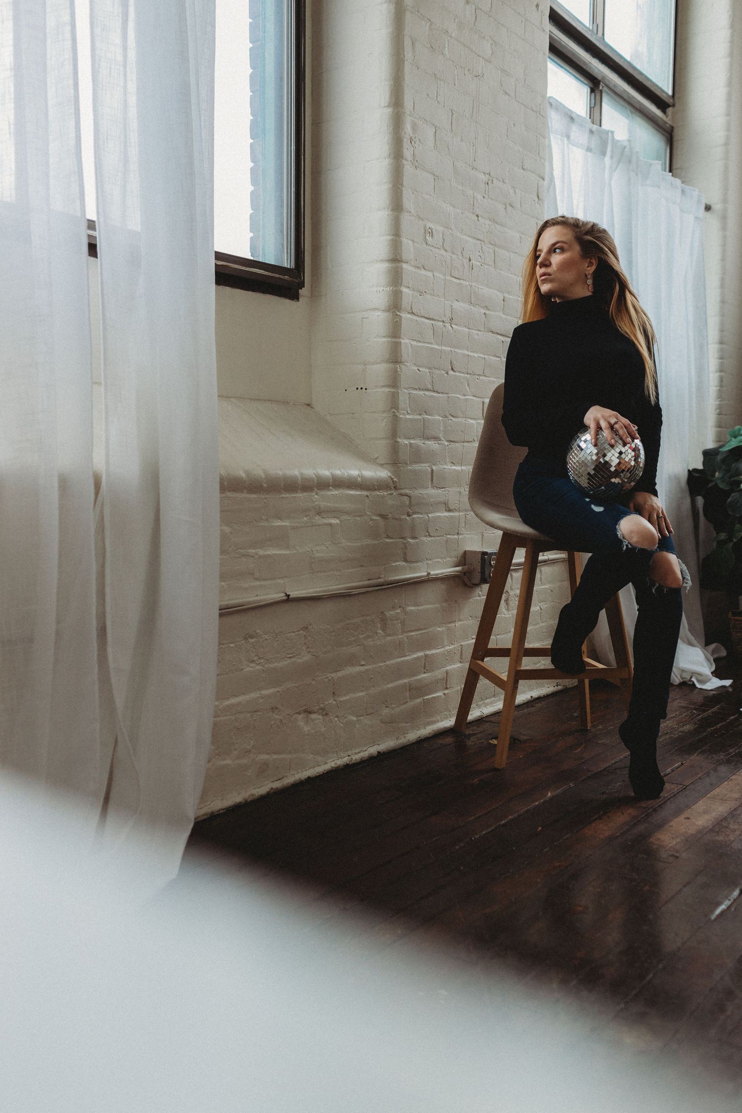 Woman in black turtleneck and stockings sits on wooden stool by white brick wall near sheer curtains.