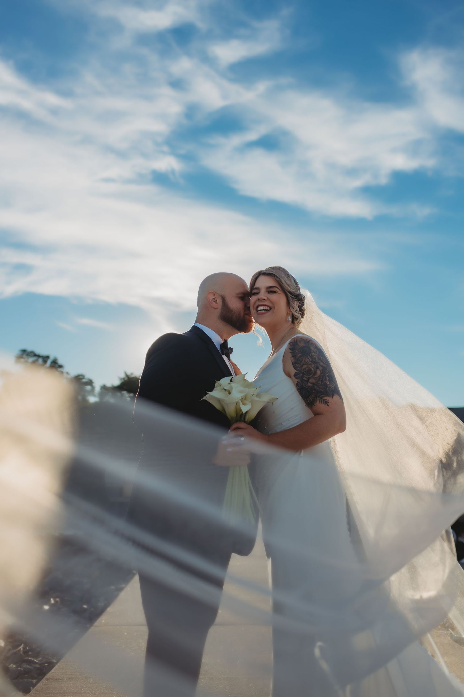 A romantic wedding couple shares a kiss against a blue sky backdrop while the bride's veil flows in the wind.