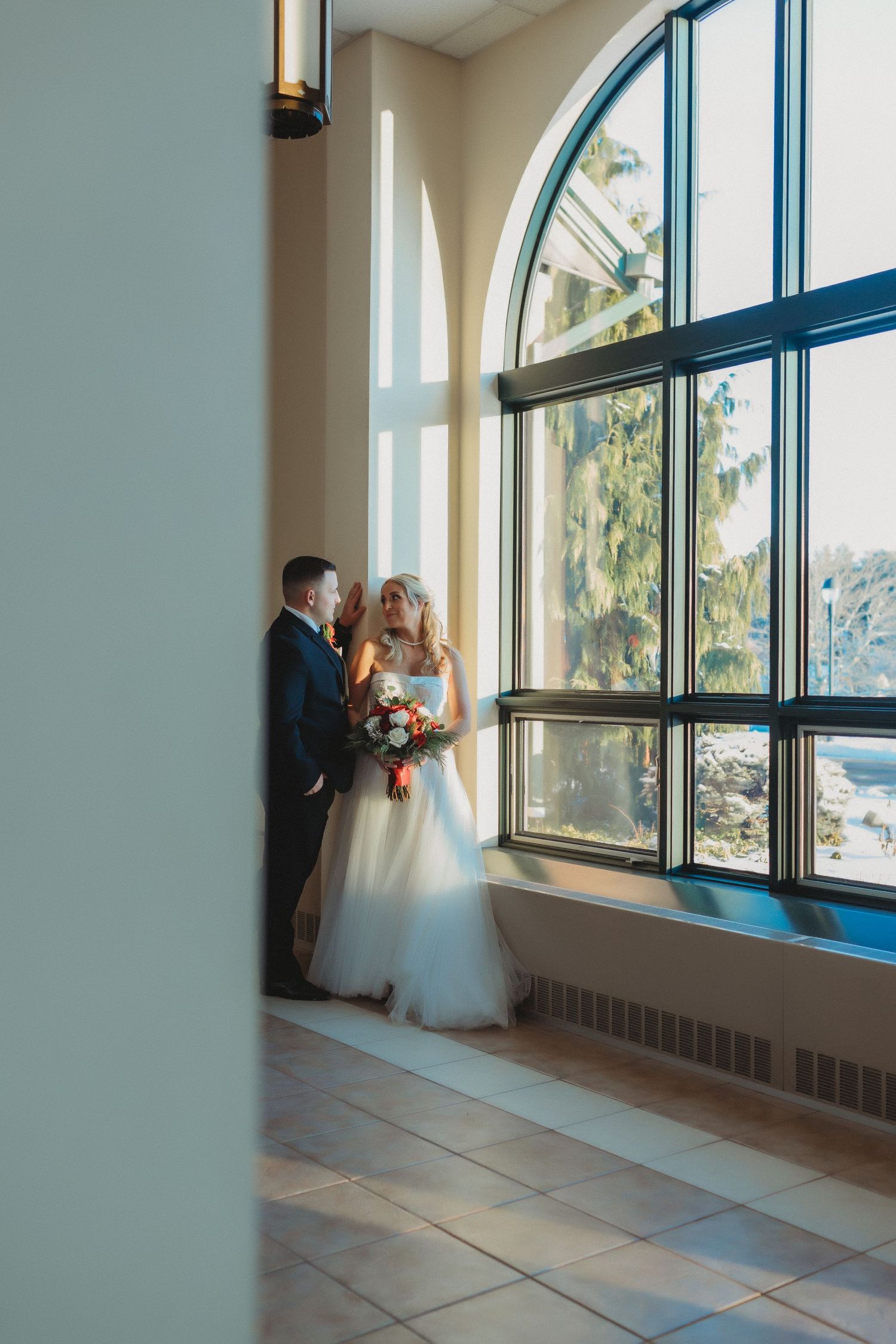 A couple in formal wedding attire stands by large arched windows in a bright interior space.