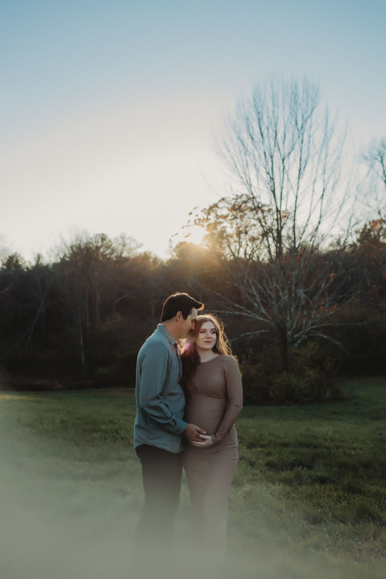 A couple embraces in a misty field at sunset with bare trees silhouetted against a soft blue sky.