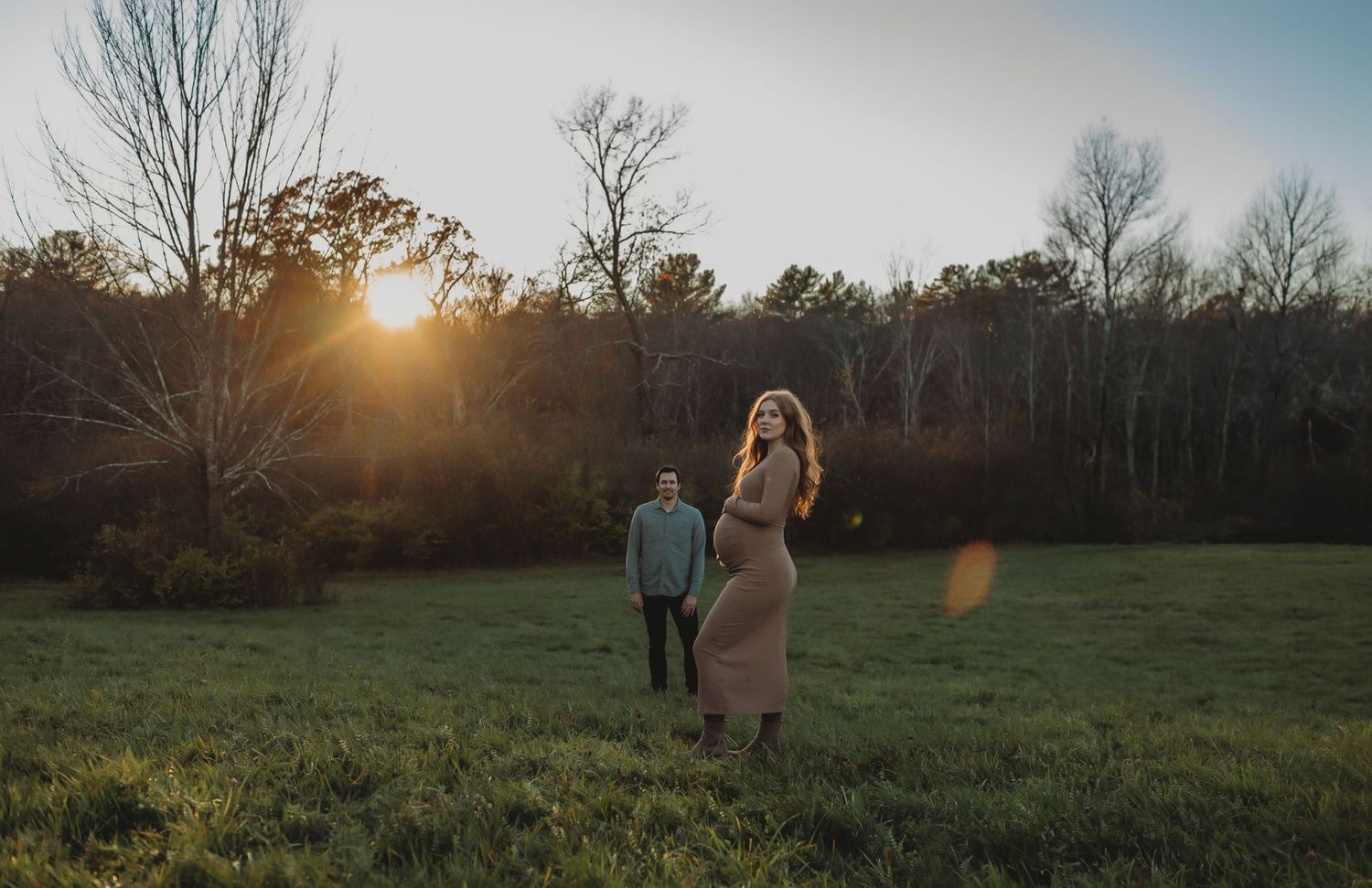 Two people stand in a grassy field at sunset with sun flare and bare trees in the background.