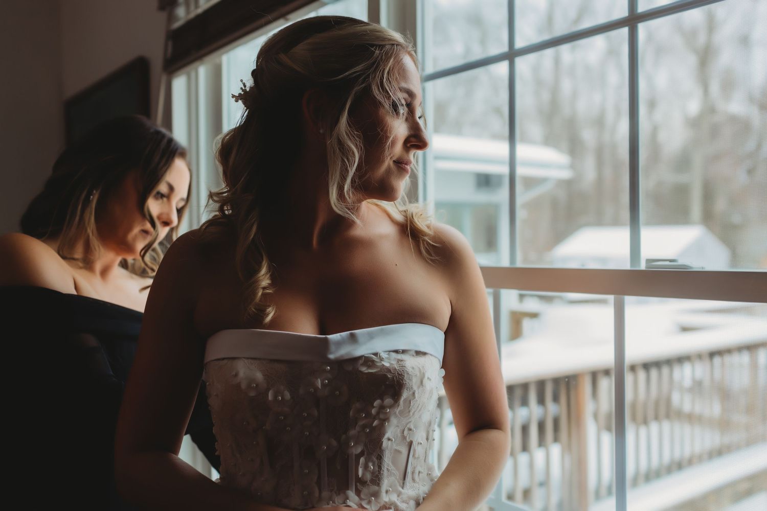 Two people stand near a window in formal attire, with one wearing an elegant white strapless gown.