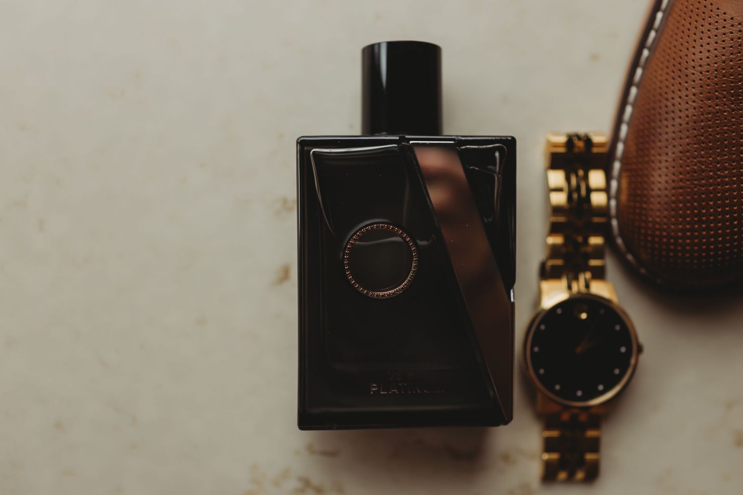 A black perfume bottle sits next to a gold watch and brown leather accessory on a light surface.