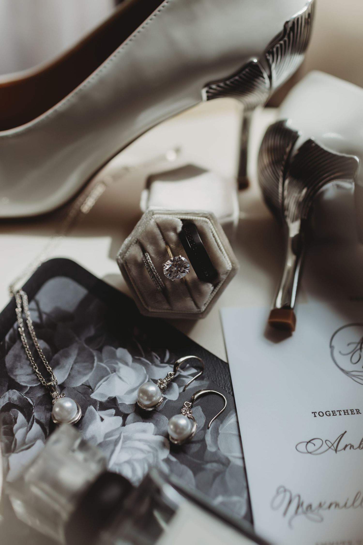 Wedding details including white heels, engagement ring in hexagonal box, pearl jewelry and invitation card on light surface.