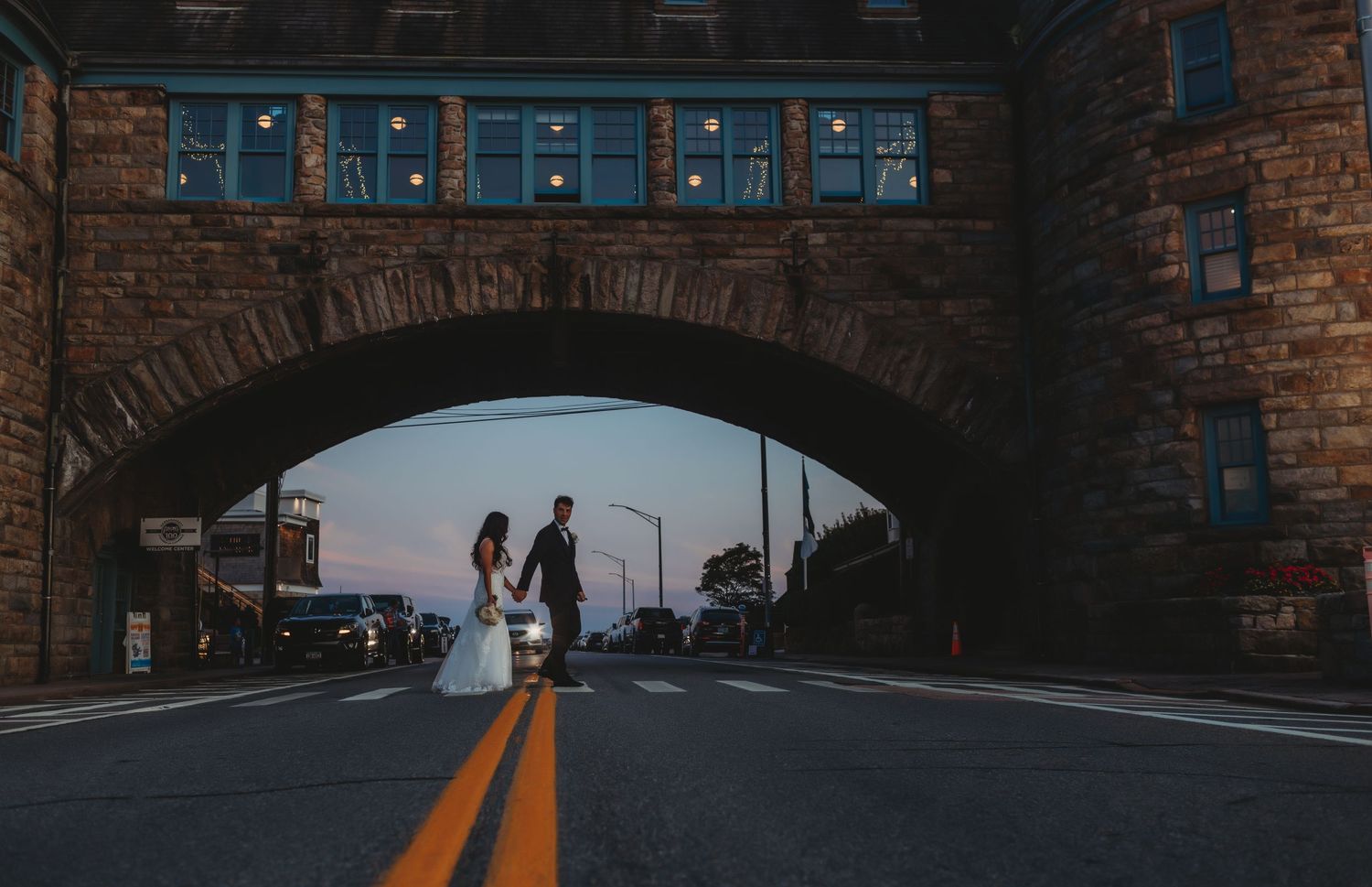 A couple silhouetted under a stone bridge at dusk with yellow road lines leading into the distance.