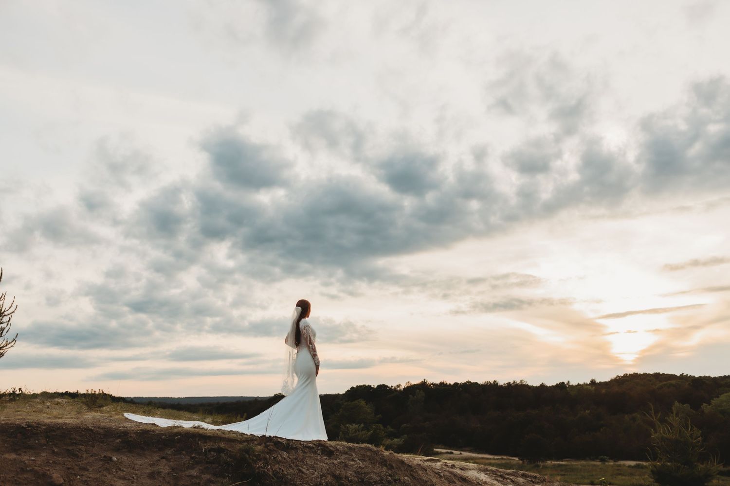 A wedding couple poses on a mountain peak against dramatic cloudy skies at sunset.