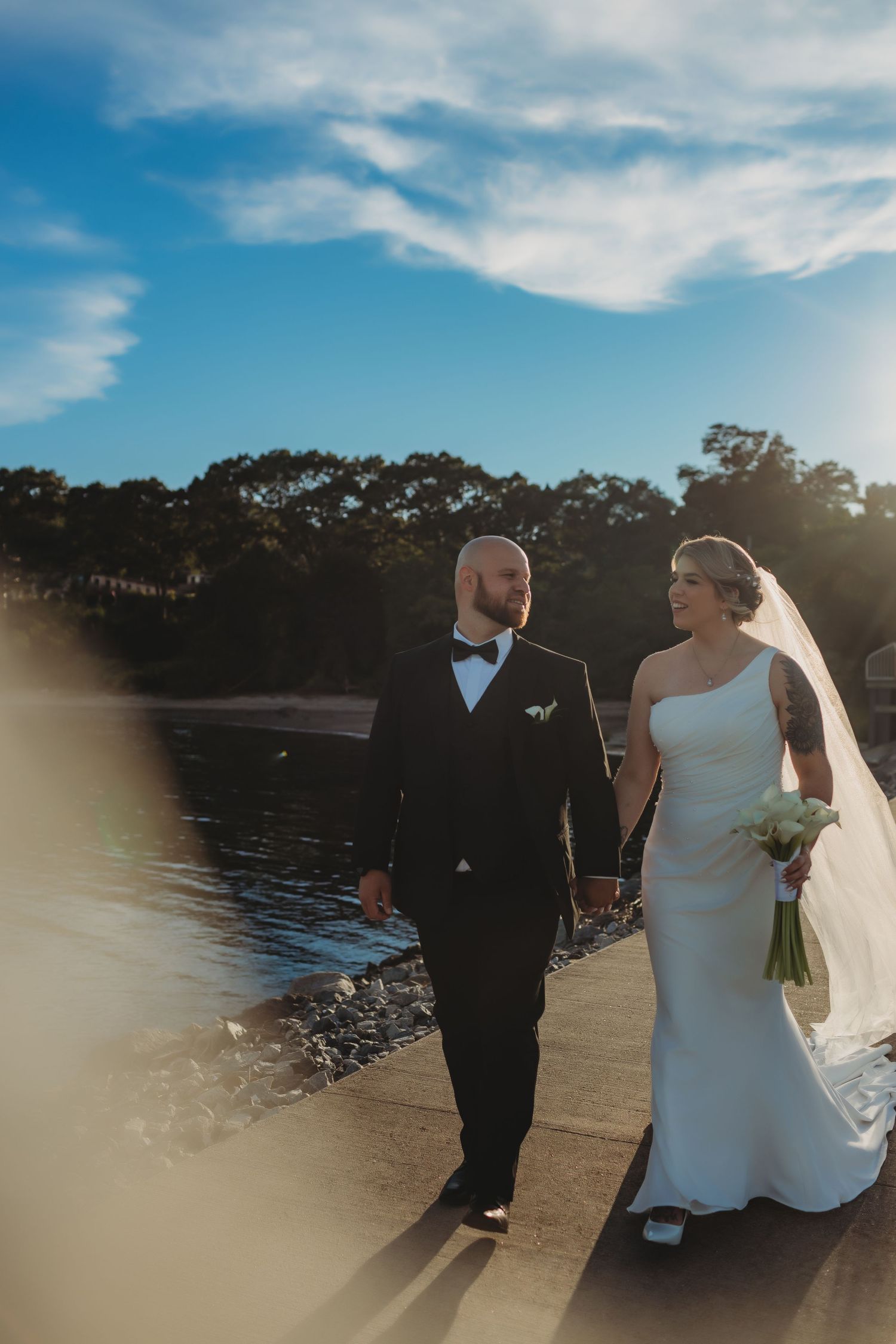 A couple in wedding attire stands together at sunset by a scenic lakeside with trees in the background.
