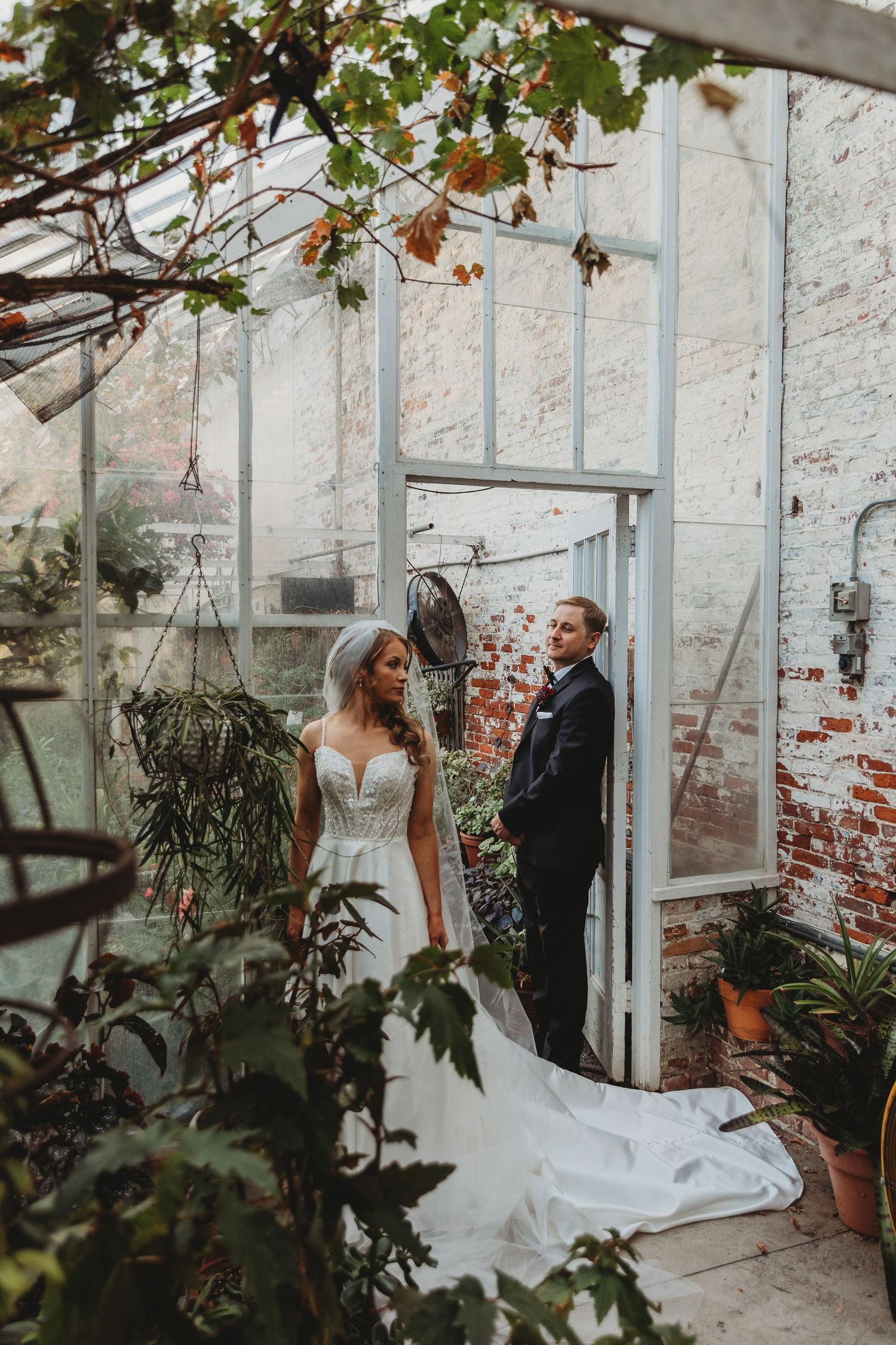 A romantic wedding photo in a rustic greenhouse with white brick walls and lush greenery framing the couple.