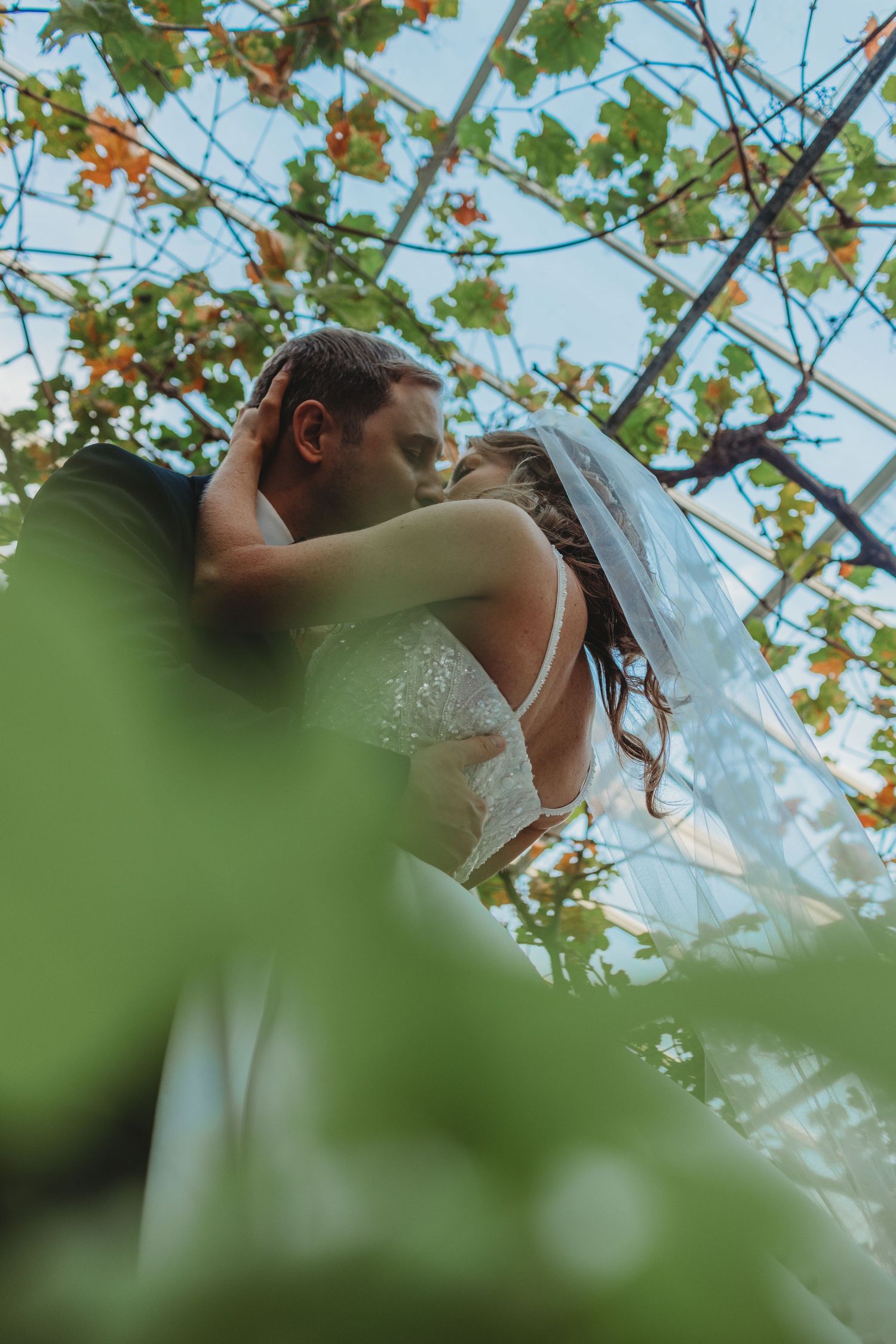 A romantic wedding couple embraces outdoors with green foliage and blue sky in the background.