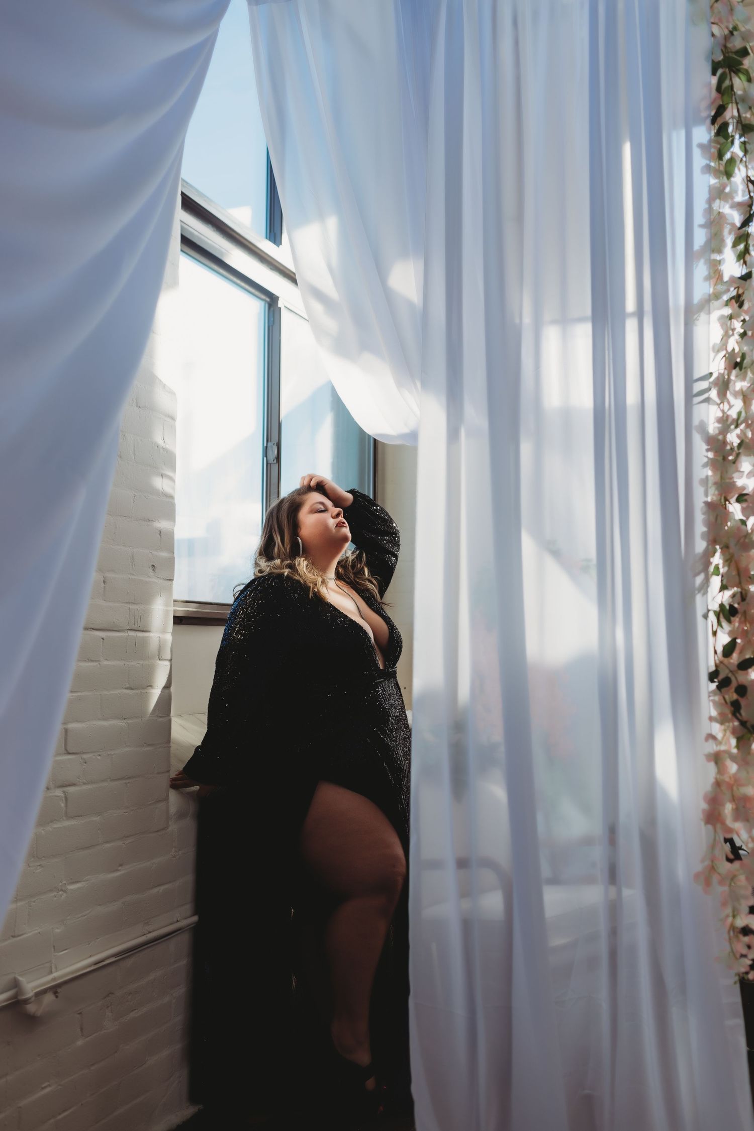 Silhouetted figure in flowing black dress poses dramatically against sheer white curtains in bright window light.