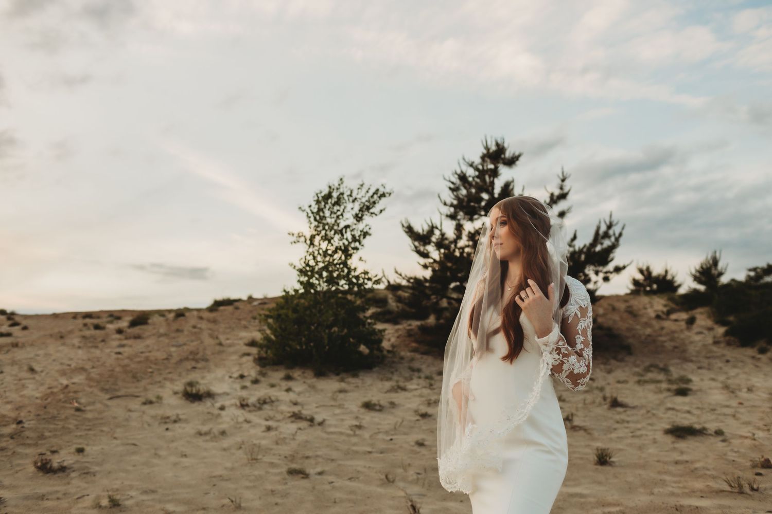 A flowing white wedding dress moves in the wind against a desert landscape with scattered trees and cloudy sky.