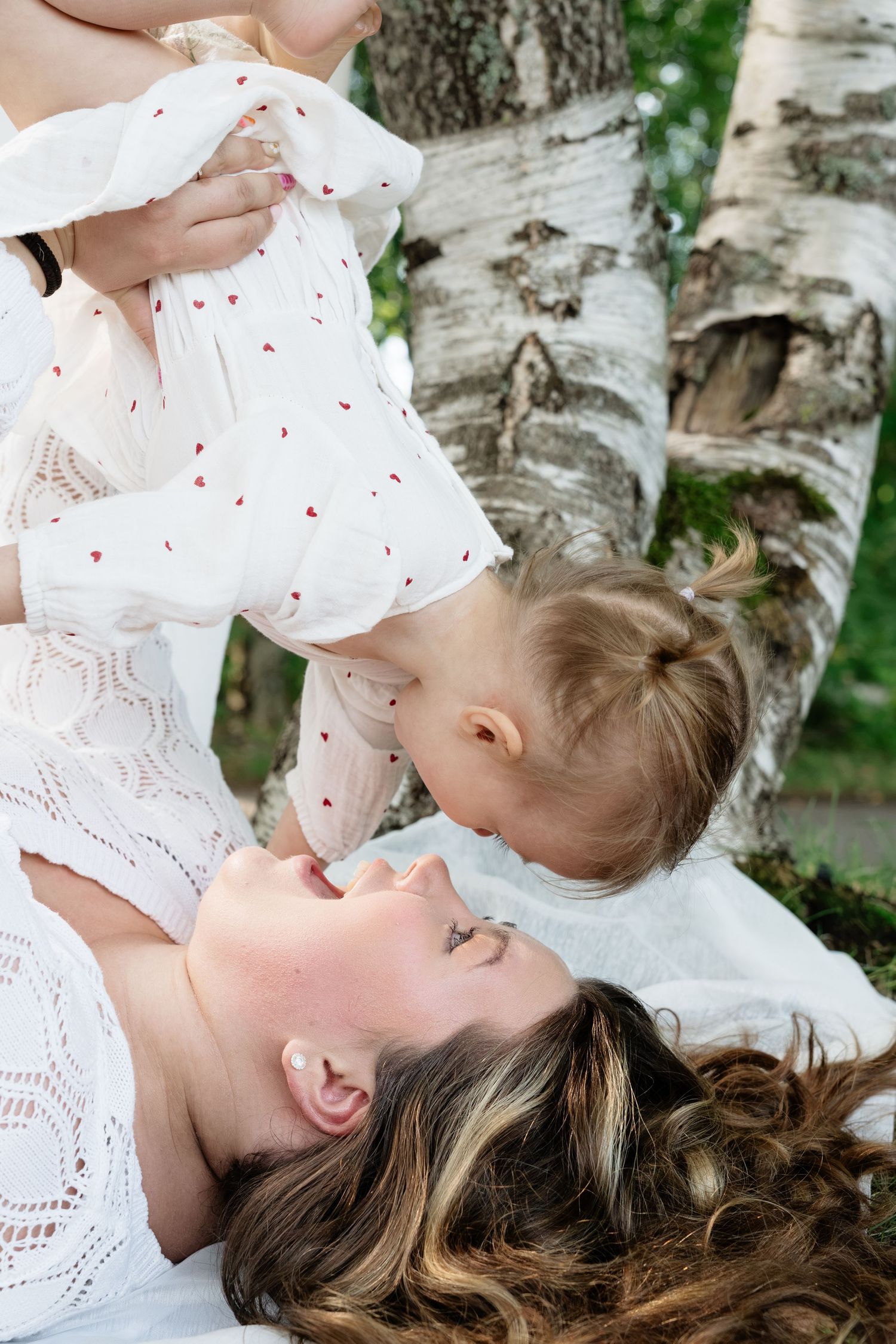Mother lying on blanket laughing as toddler daughter in white heart dress leans in for a kiss outdoors near birch trees.