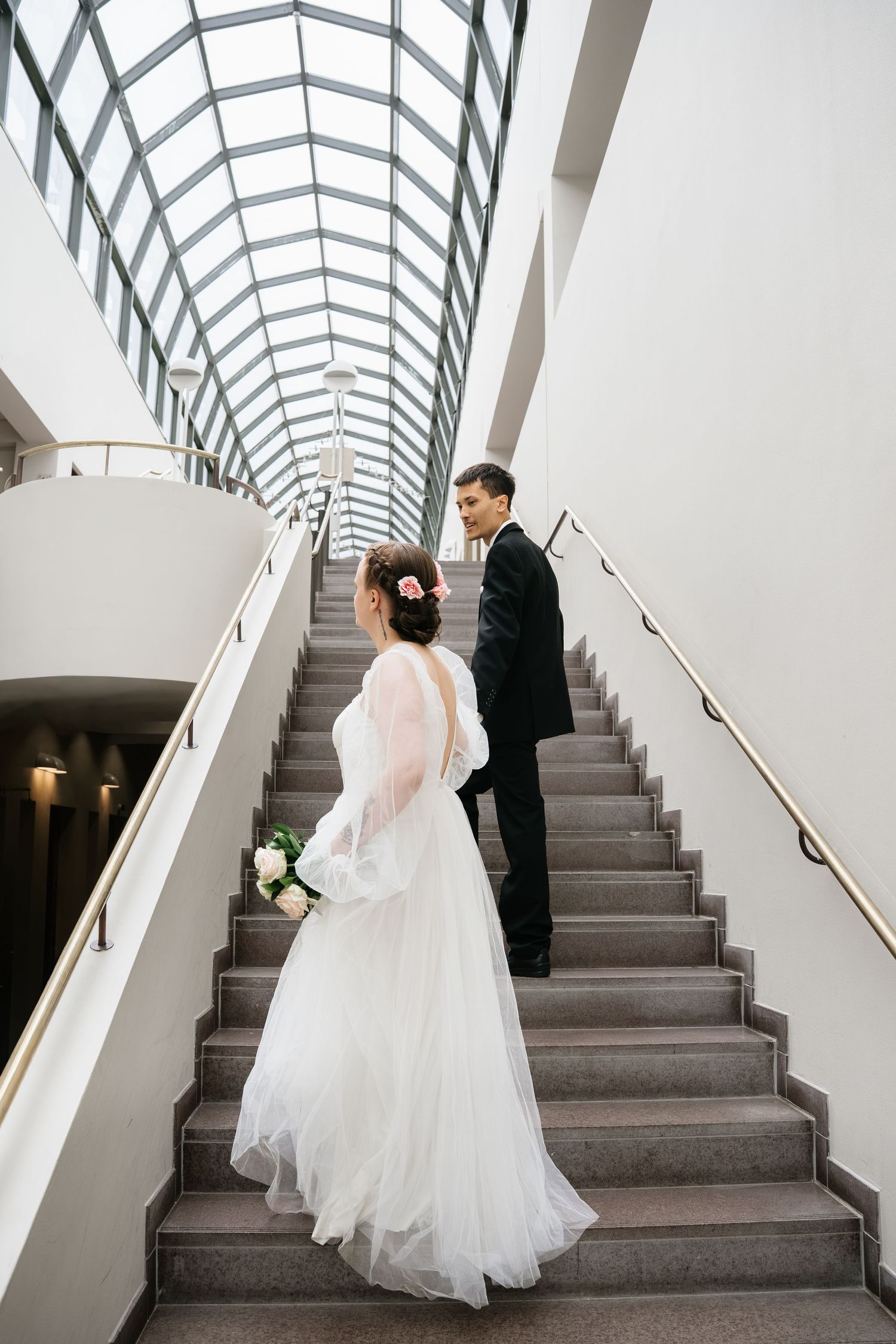 Bride in white gown and groom in black suit walking up elegant staircase with glass ceiling at wedding venue.
