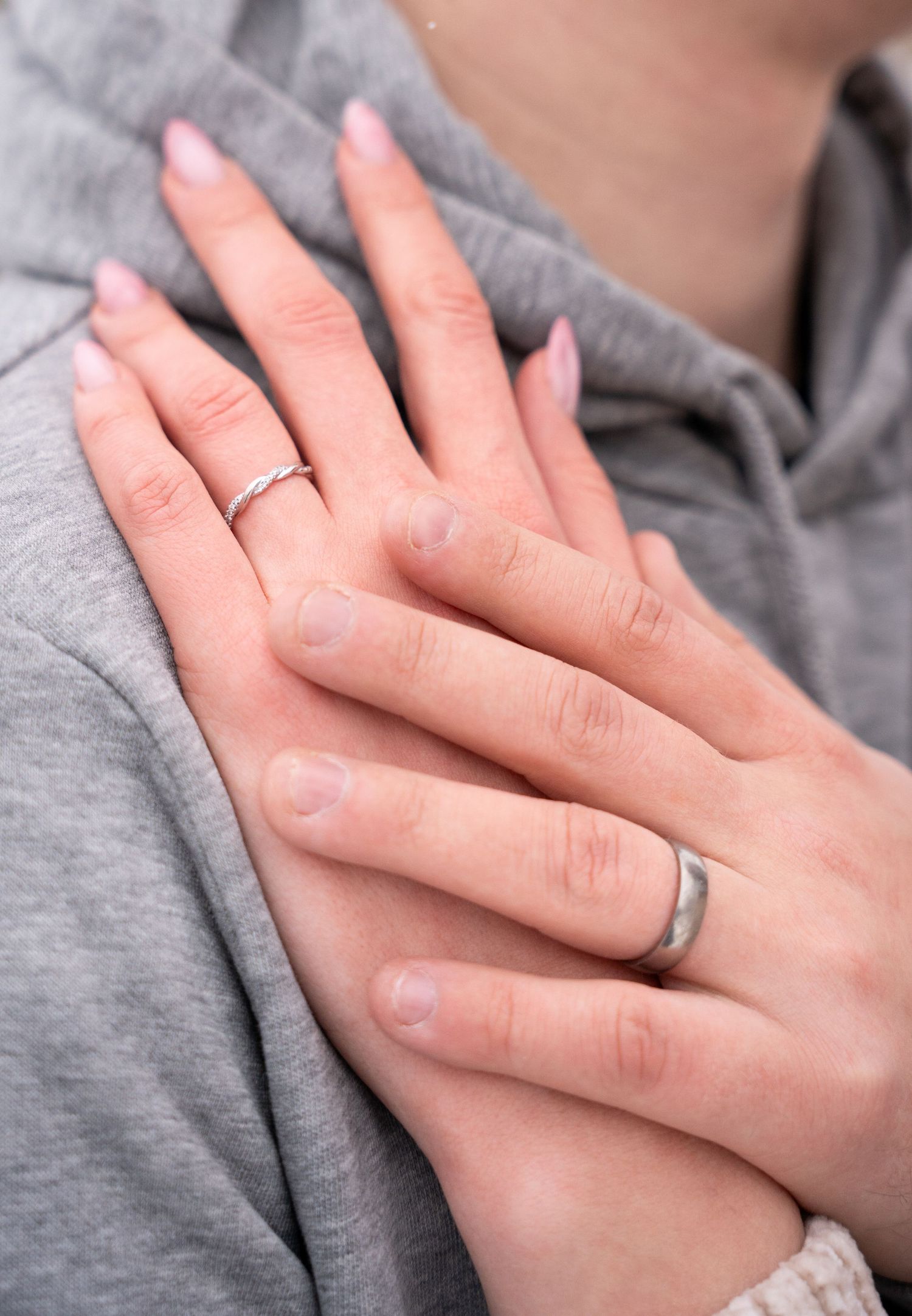 Close-up of couple's hands showing matching wedding bands, woman with pink manicure resting on man's chest in gray hoodie.
