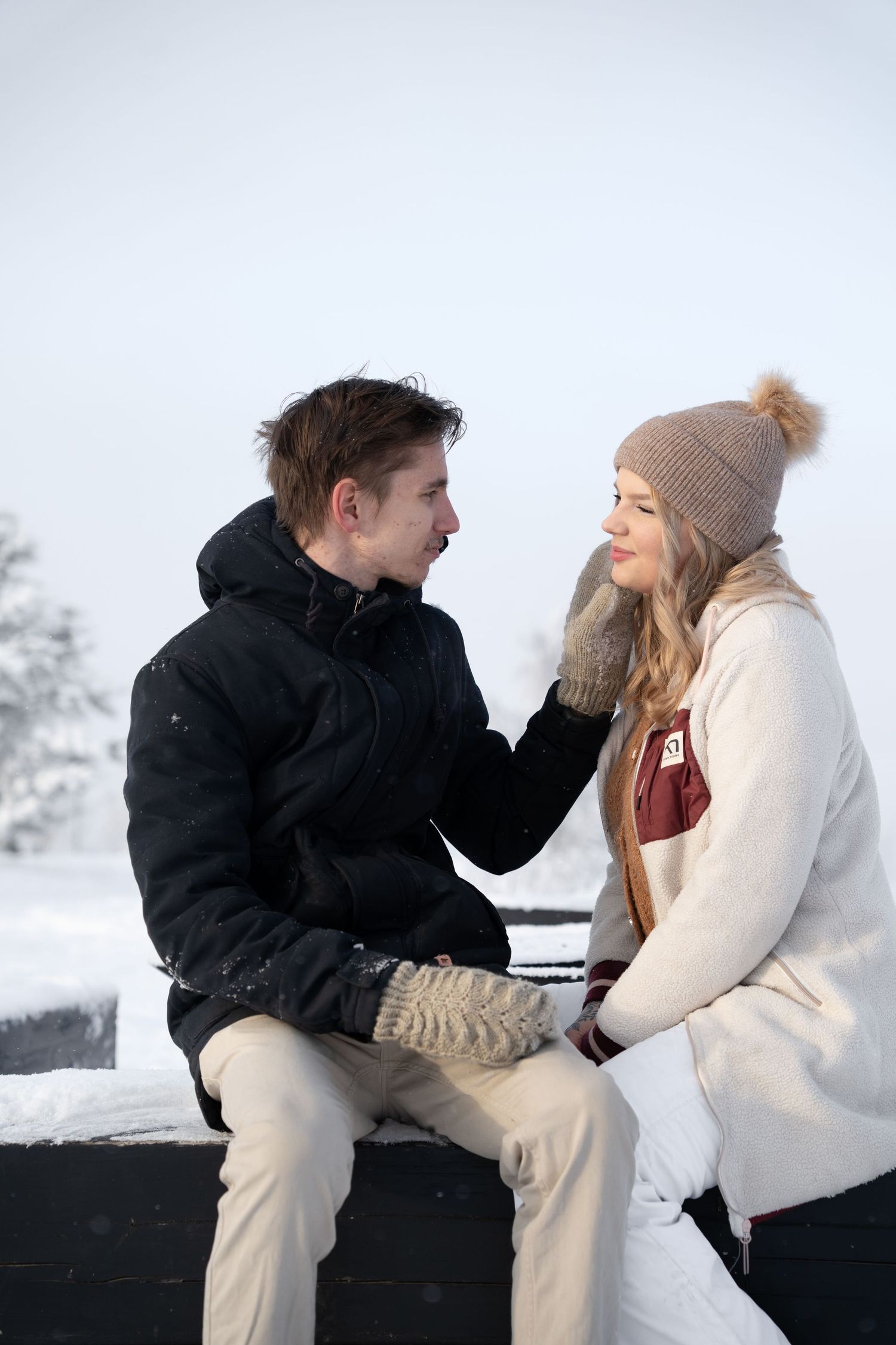 Young couple sitting together on snowy surface, smiling at each other, woman in pom-pom beanie touching man's face gently.