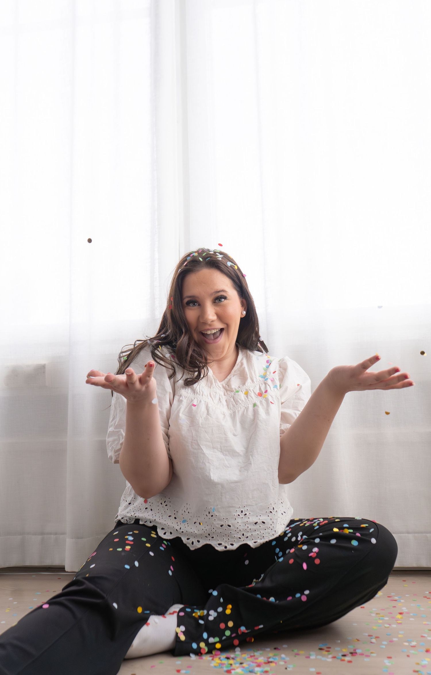 Happy woman sitting cross-legged on floor tossing colorful confetti with joyful expression indoors.